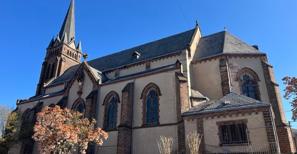 Seitlicher Blick auf eine Kirch mit spitzem Turm, die auf einem kleinen grünen Hang steht; im Vordergrund ein blühender Baum.
