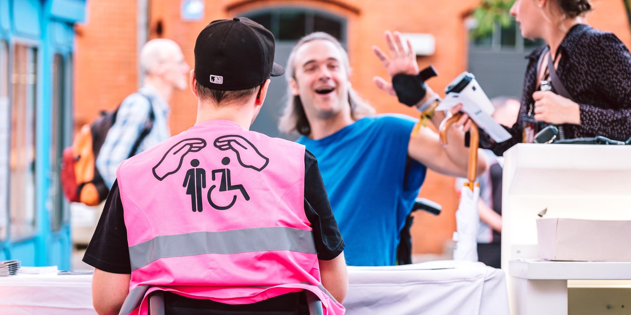 Links im Bild sieht man den Rücken eines Rollstuhlfahrers mit einer pinken Weste. Darauf abgebildet sind zwei Hände, schützend über zwei Symbolen: links das für All gender welcome und rechts das für Rollstuhlfahrer*innen. Im Hintergrund sitzt ein junger Mann in blauem T-Shirt, ebenfalls Rollstuhlfahrer, der in die Kamera winkt und lächelt.
