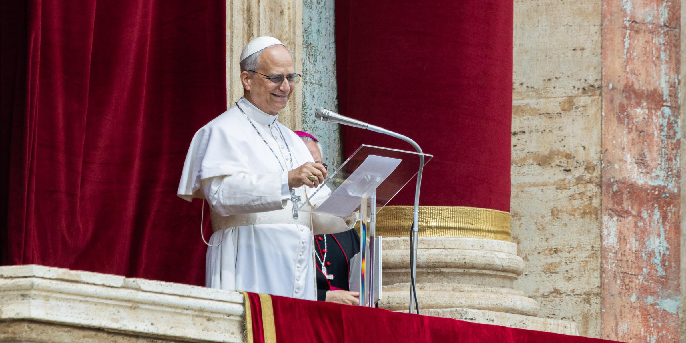 Papst Leo XIV. steht in weißer päpstlicher Kleidung auf der Loggia des Petersdoms vor einem roten Samtvorhang und spricht an einem Mikrofon. Er trägt ein Brustkreuz und eine weiße Kappe (Zucchetto) und lächelt. Eine weitere geistliche Person in dunkler Kleidung ist teilweise hinter ihm zu sehen.