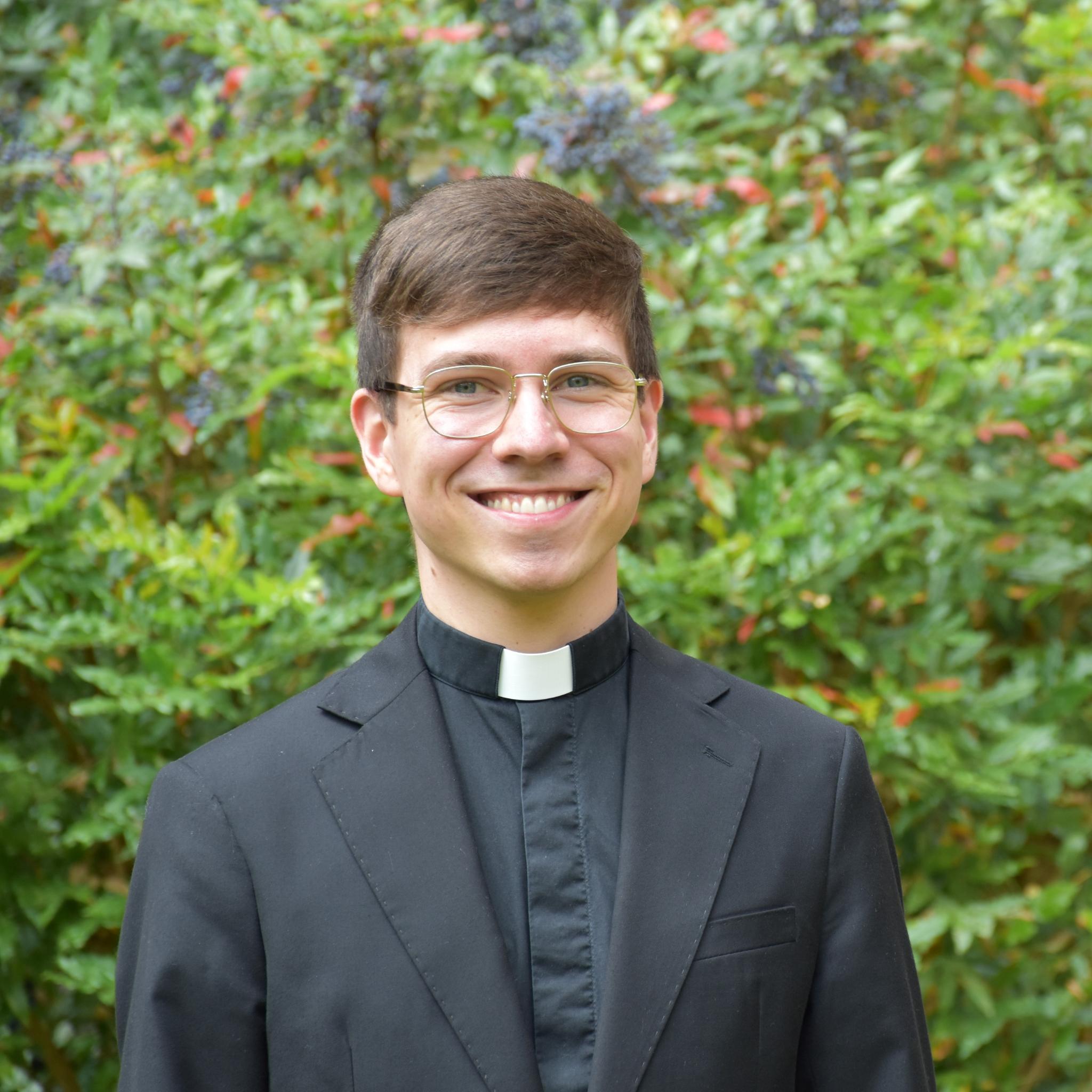 Junger Mann mit Brille, braunem kurzem Haar, schwarzem Anzug und Priesterkragen steht lächelnd vor einem grünen Strauch