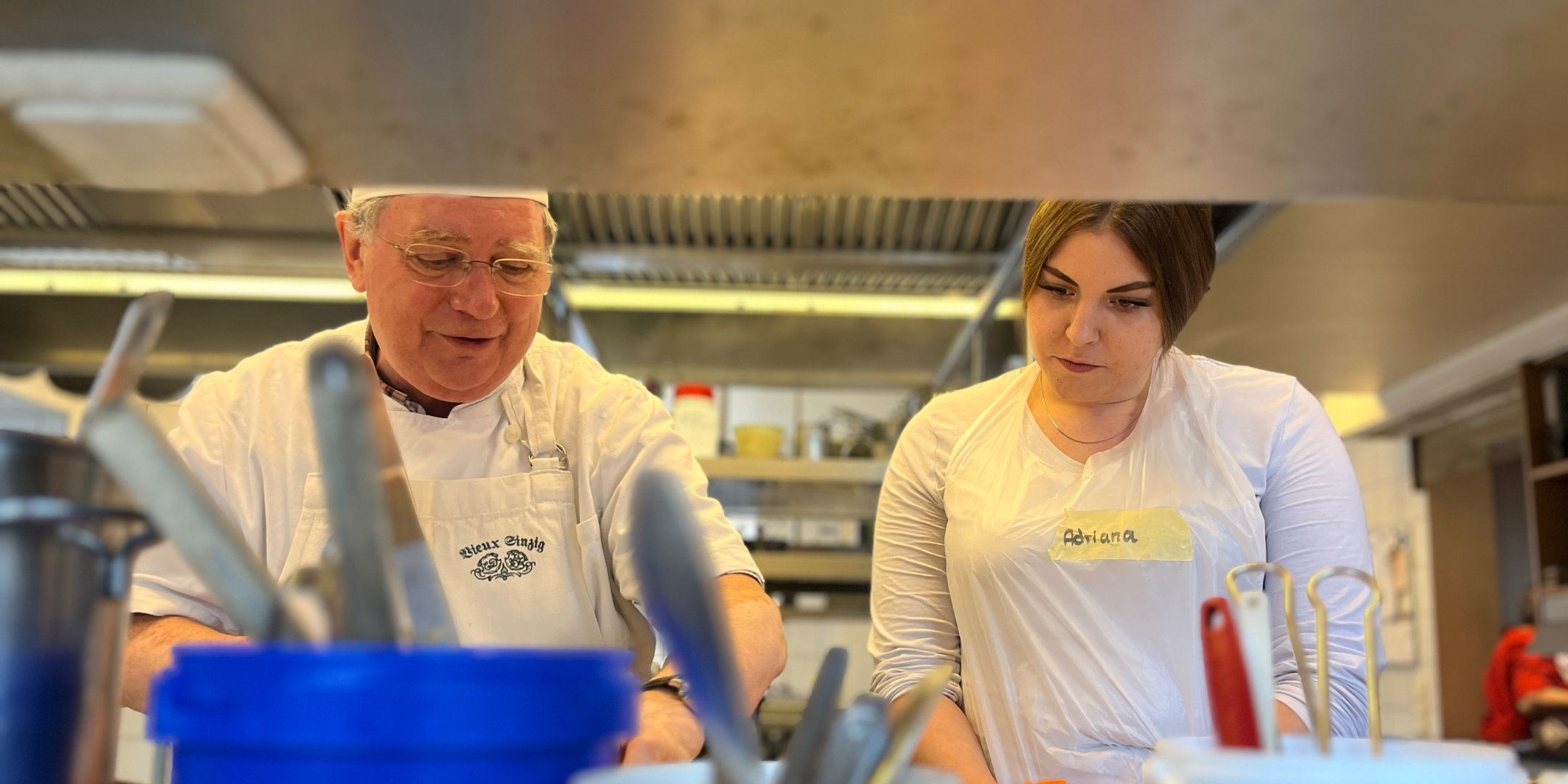 Ein Koch steht mit Kochmütze in einer Küche, neben ihm eine junge Frau, die ihm zuschaut.