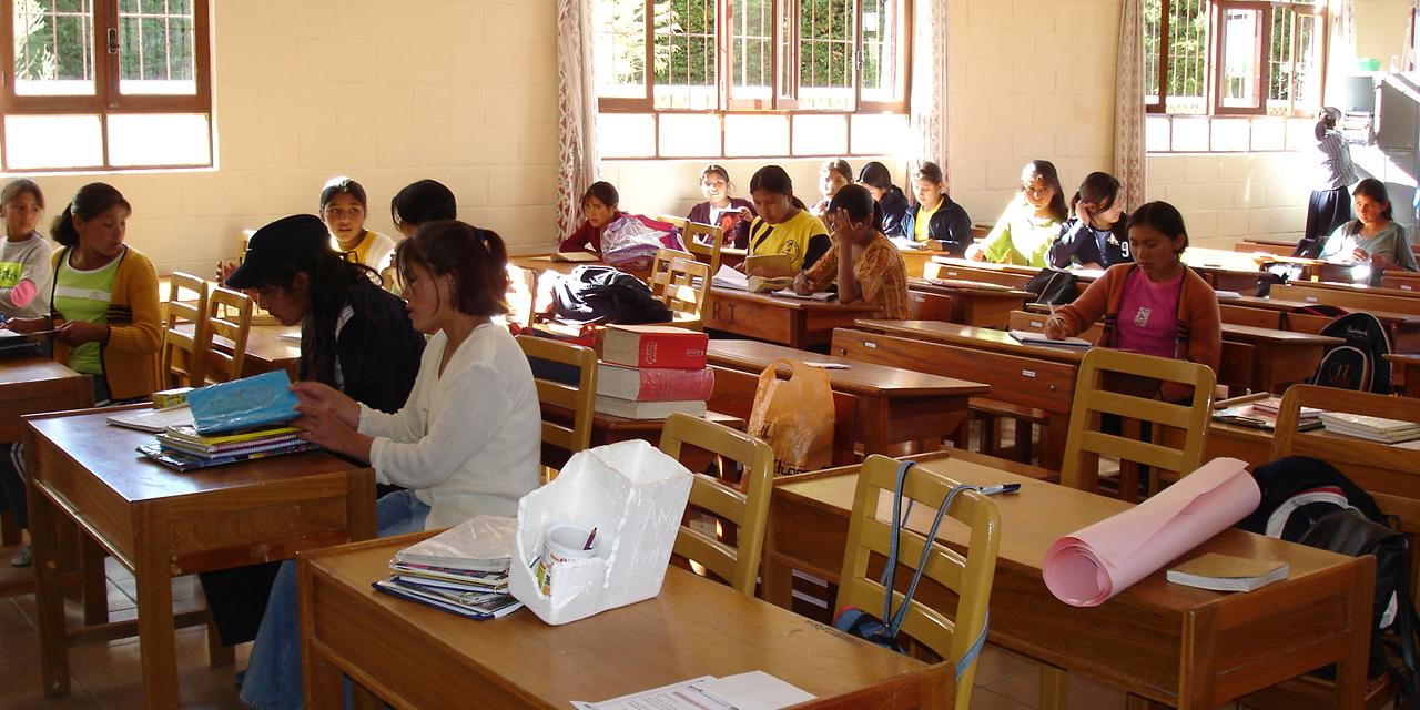 Das Foto zeigt einen Klassenraum in Bolivien. Holztische mit jeweils zwei Stühlen stehen in drei Reihen nebeneinander. Auf den Tischen in der vorderen Reihen liegen Bücher und Papier, hier sitzen keine Schülerinnen und Schüler. Die Tische in der mittleren und hinteren Reihe sind mit jeweils ein oder zwei Schülerinnen besetzt, die schreiben, lesen und sich unterhalten. Die hintere Reihe steht vor einer Wand mit Fenstern, draußen scheint die Sonne, vor den Fenstern stehen Büsche oder eine Hecke.