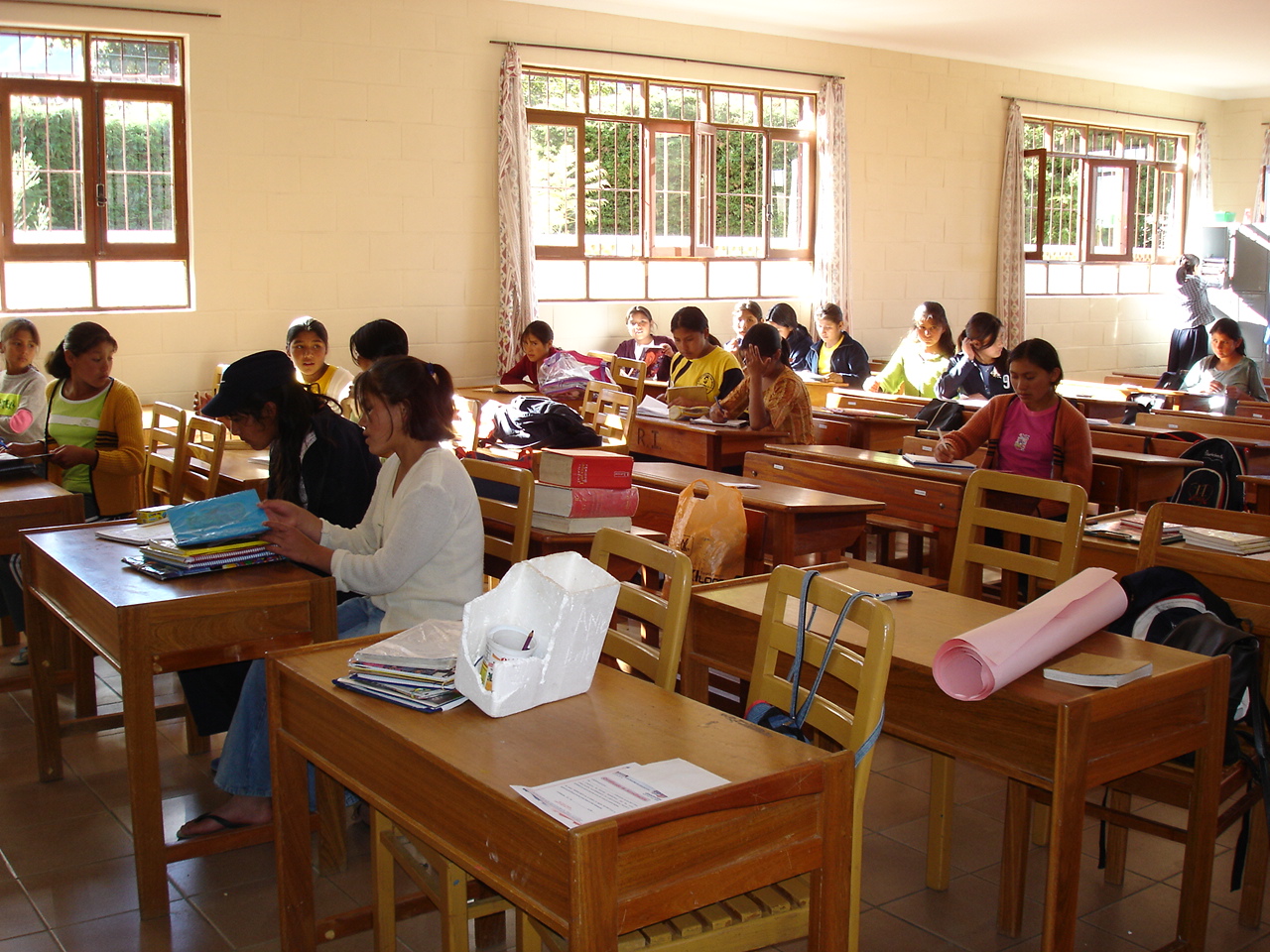 Das Foto zeigt einen Klassenraum in Bolivien. Holztische mit jeweils zwei Stühlen stehen in drei Reihen nebeneinander. Auf den Tischen in der vorderen Reihen liegen Bücher und Papier, hier sitzen keine Schülerinnen und Schüler. Die Tische in der mittleren und hinteren Reihe sind mit jeweils ein oder zwei Schülerinnen besetzt, die schreiben, lesen und sich unterhalten. Die hintere Reihe steht vor einer Wand mit Fenstern, draußen scheint die Sonne, vor den Fenstern stehen Büsche oder eine Hecke.