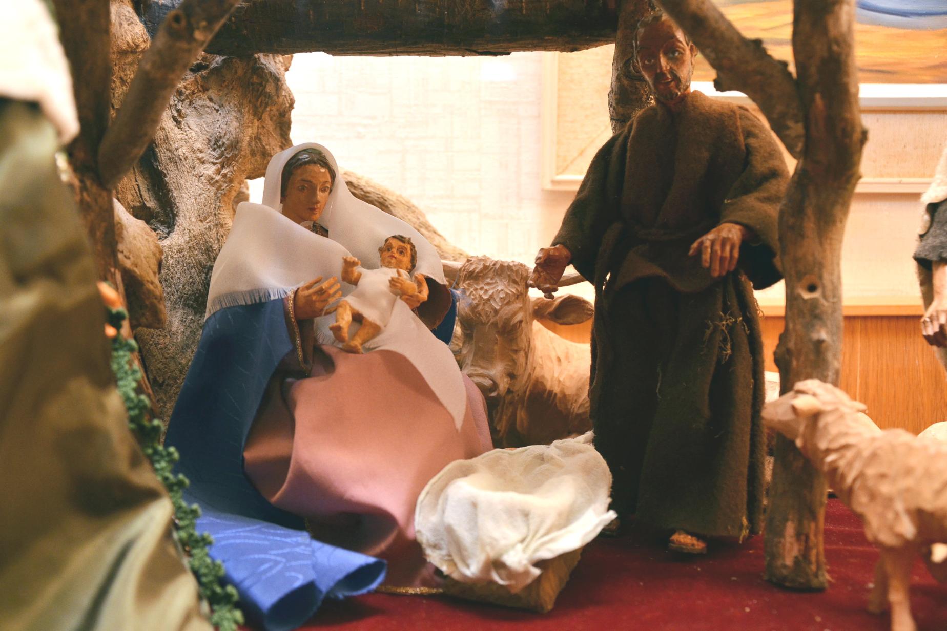 Das Foto zeigt Krippenfiguren. Links sitzt Maria, sie hält den neugeborenen Jesus auf dem Arm. Rechts steht Josef. Zwischen den beiden am Boden die leere Futterkrippe, dahinter der Ochse. Von rchts schaut ein Schaf in die Szene.