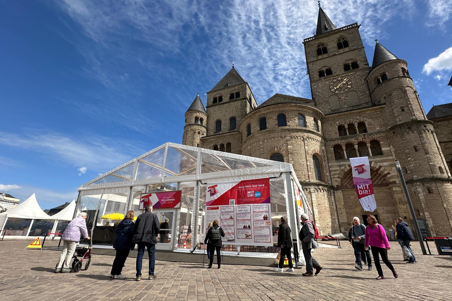 Man sieht das Kulturzelt vor dem Dom, der Himmel ist blau; im Vordegrund stehen einige Menschen auf dem Domfreihof-