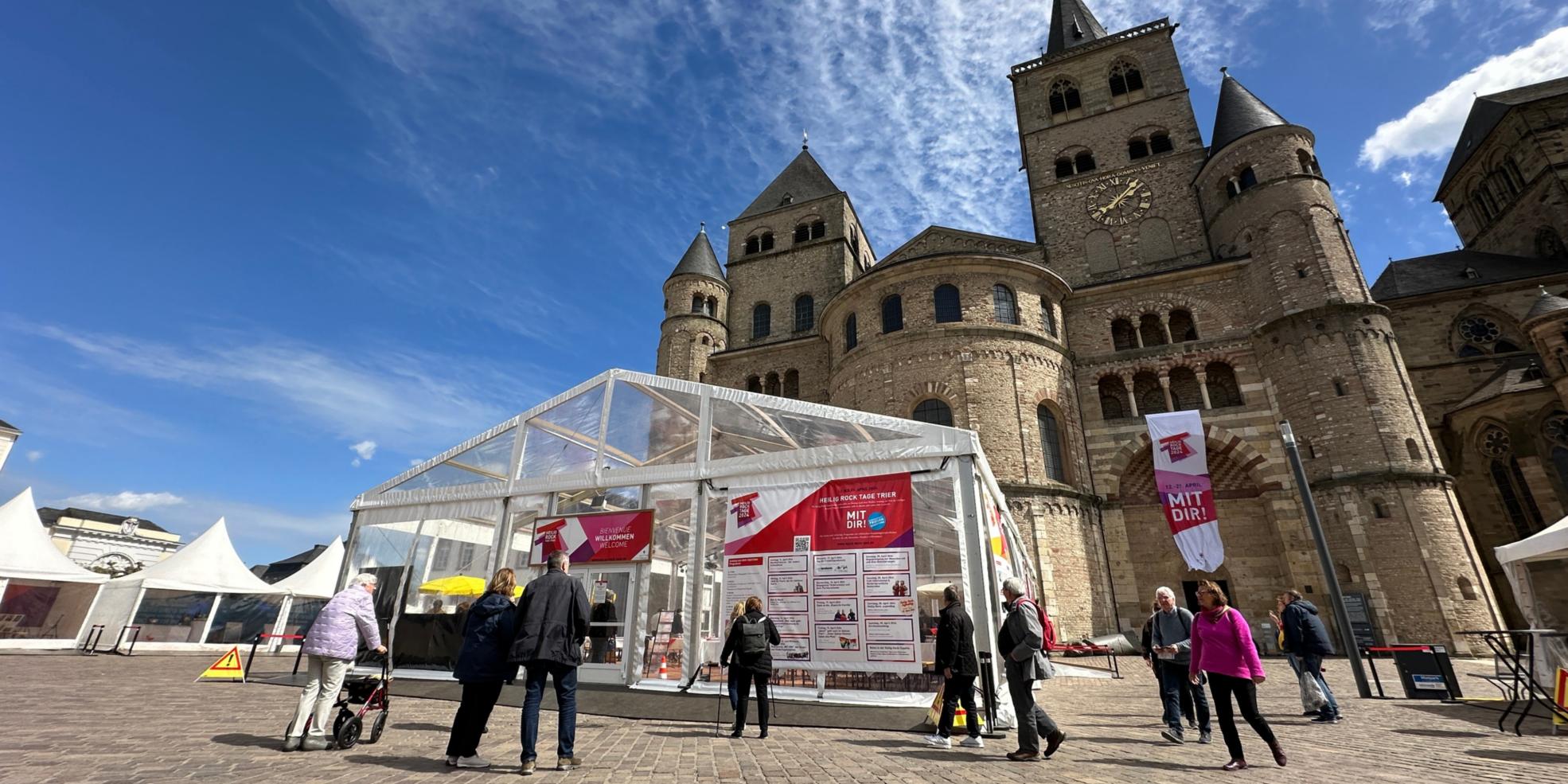 Man sieht das Kulturzelt vor dem Dom, der Himmel ist blau; im Vordegrund stehen einige Menschen auf dem Domfreihof-