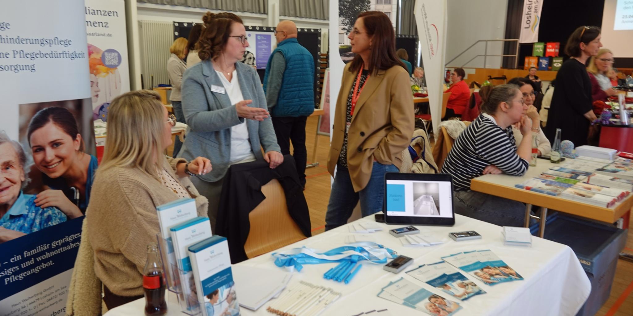 In einem großen Saal mit mehreren Informationsständen sitzen und stehen verschiedene Ausstellerinnen und Besucher an Tischen. Im Vordergrund befindet sich ein Stand mit Broschüren, Flyern, Stiften, einem Tablet mit geöffneter Präsentation sowie medizinischem Informationsmaterial. Hinter dem Tisch sprechen mehrere Personen miteinander. Weitere Stände mit Plakaten, Roll-ups und Infomaterial sind im Hintergrund zu sehen. Der Raum ist hell, mit großen Fenstern, Bühnenbereich und zahlreichen Besucherinnen und Besuchern, die sich austauschen und informieren.