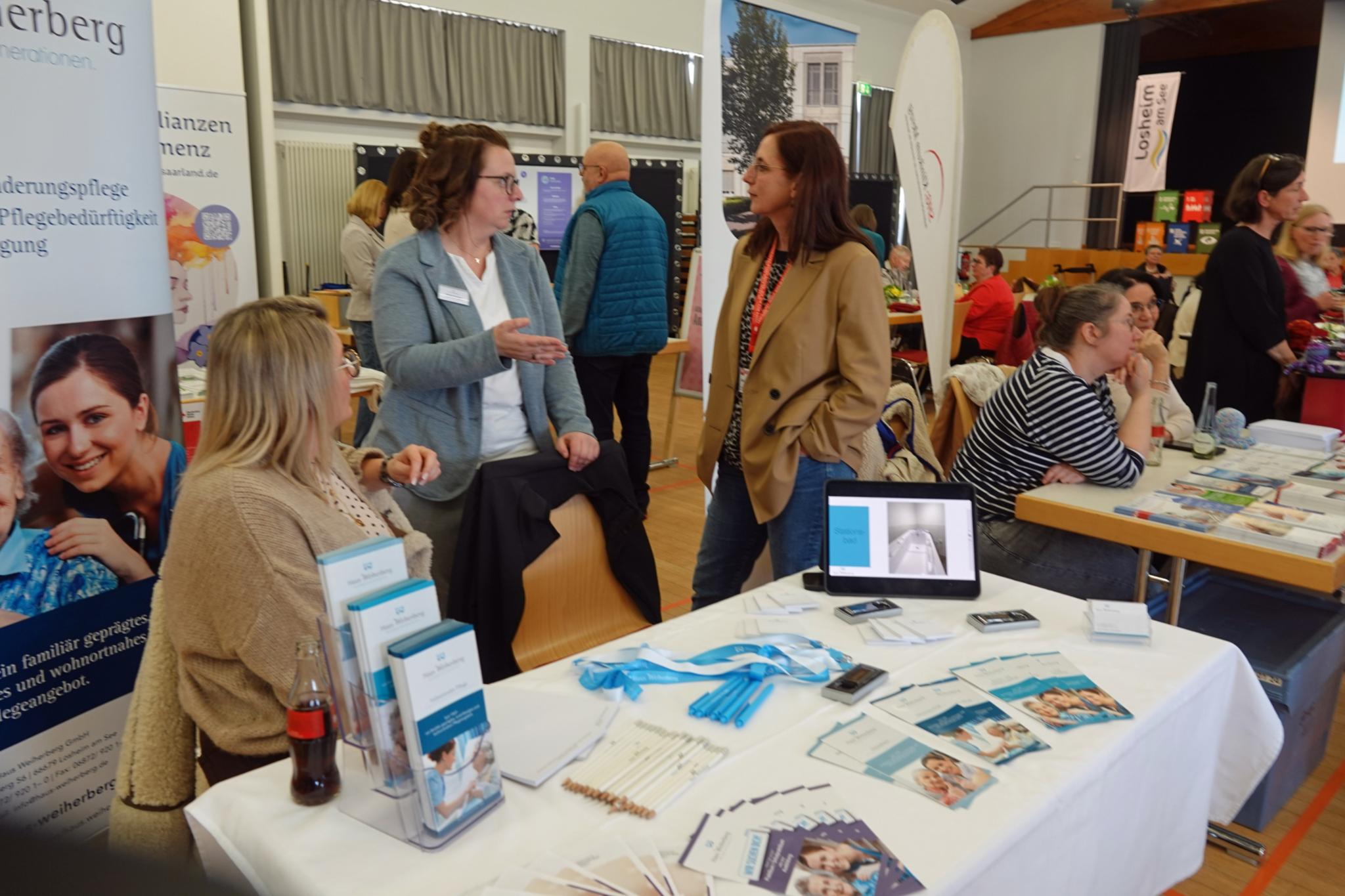 In einem großen Saal mit mehreren Informationsständen sitzen und stehen verschiedene Ausstellerinnen und Besucher an Tischen. Im Vordergrund befindet sich ein Stand mit Broschüren, Flyern, Stiften, einem Tablet mit geöffneter Präsentation sowie medizinischem Informationsmaterial. Hinter dem Tisch sprechen mehrere Personen miteinander. Weitere Stände mit Plakaten, Roll-ups und Infomaterial sind im Hintergrund zu sehen. Der Raum ist hell, mit großen Fenstern, Bühnenbereich und zahlreichen Besucherinnen und Besuchern, die sich austauschen und informieren.