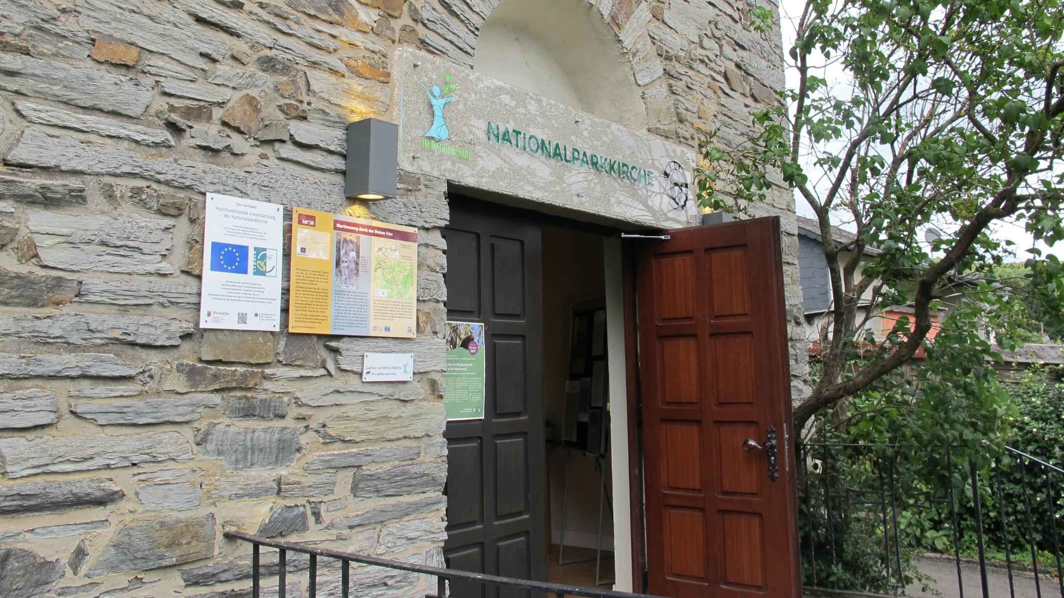 Gebäude mit alter Steinmauer mit offenen Holztür im Grünen, darüber hängt ein Schild mit der Aufschrift Nationalparkkirche