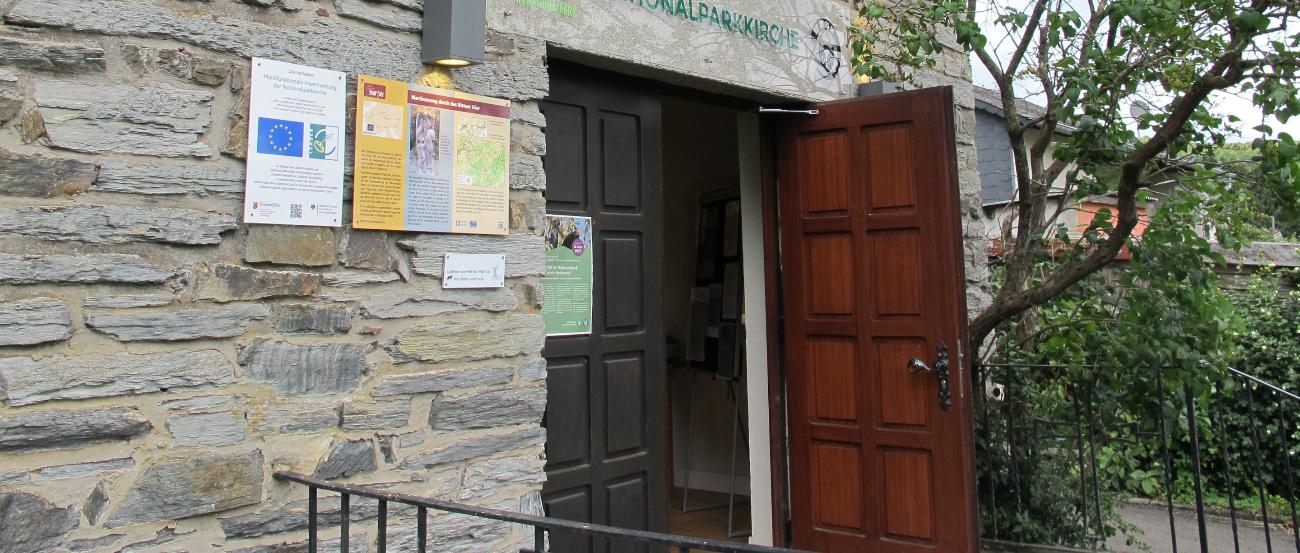 Gebäude mit alter Steinmauer mit offenen Holztür im Grünen, darüber hängt ein Schild mit der Aufschrift Nationalparkkirche