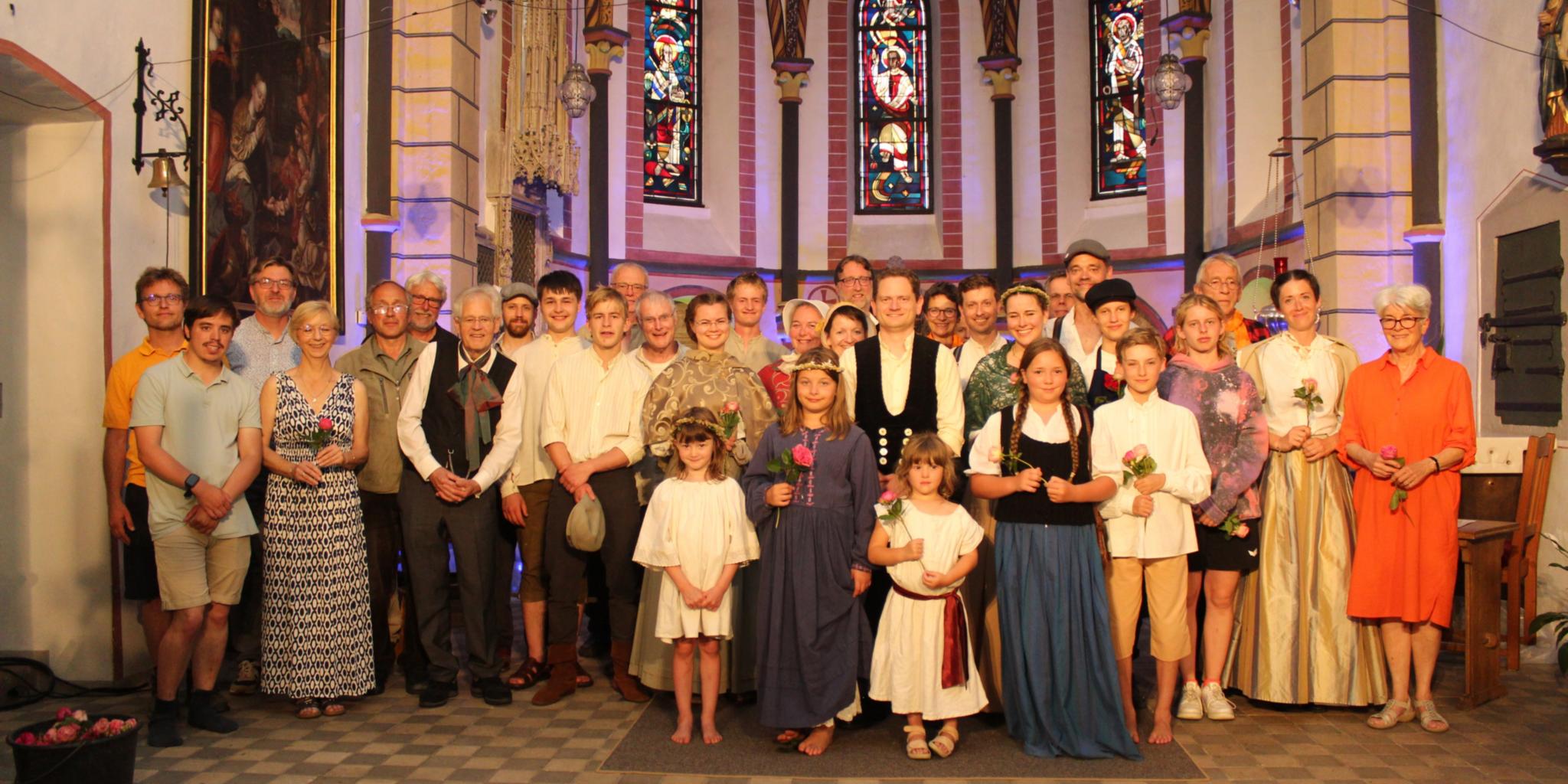 Eine große Gruppe von Menschen steht gemeinsam im Altarraum einer Kirche. Viele tragen historische oder festliche Kleidung, einige moderne Kleidung. Kinder und Erwachsene stehen gemischt in mehreren Reihen. Hinter ihnen sind hohe, bunt leuchtende Kirchenfenster und gotische Architektur zu erkennen.