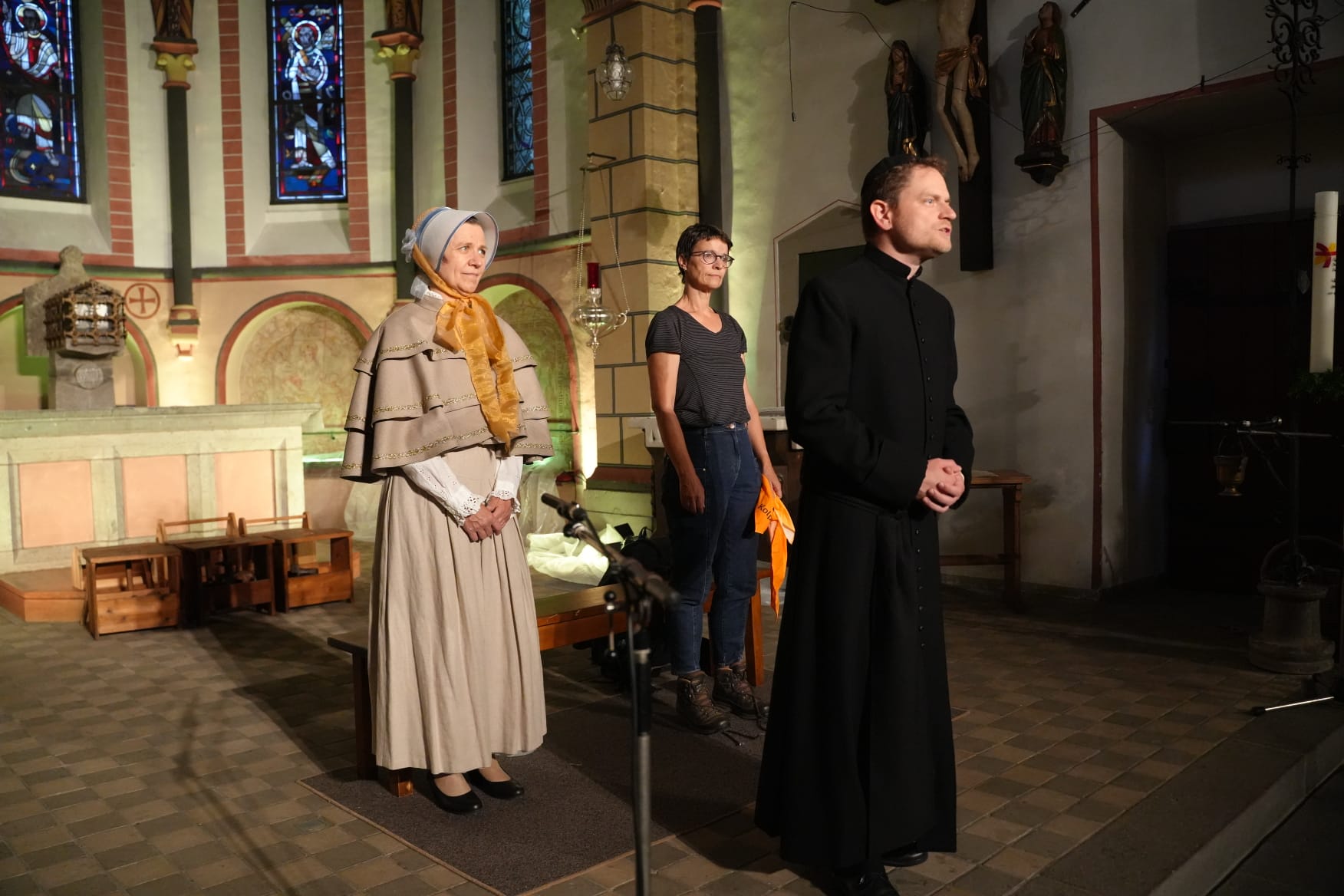 Eine Szene in einer Kirche: Drei Personen stehen vorne im Altarraum. Links eine Person in historischer Kleidung mit langem Rock, Kopftuch und Umhang; in der Mitte eine Person in moderner Alltagskleidung mit Jeans und T‑Shirt; rechts eine Person in einem langen schwarzen Gewand, ähnlich einer priesterlichen Robe. Hinter ihnen sind Kirchenbänke, ein Altar und farbige Kirchenfenster zu sehen.