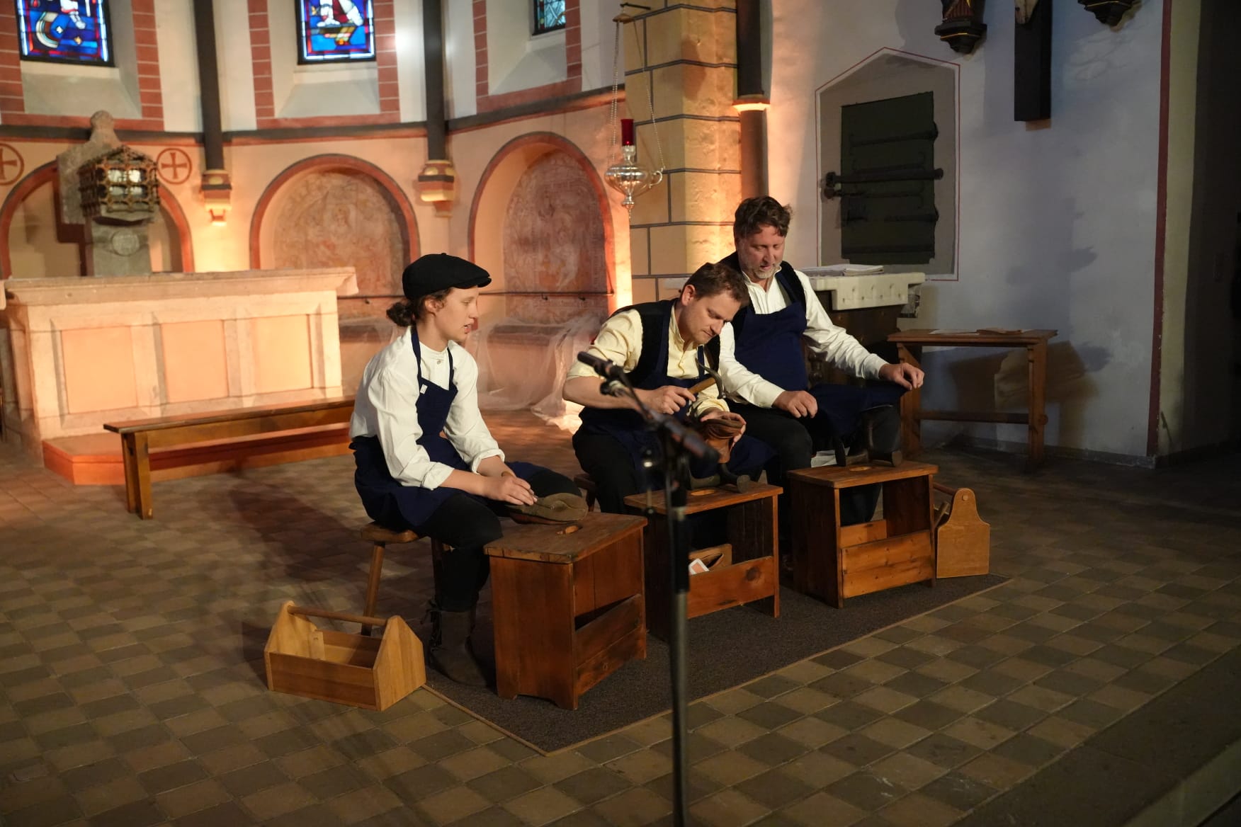 Drei Personen in traditioneller Handwerkskleidung sitzen in der Kirche auf kleinen Holzbänken und arbeiten an Holzschuhen oder Werkzeugen. Sie tragen weiße Hemden, dunkle Hosen und Schürzen. Vor ihnen stehen Holzkisten mit Werkzeugen. Der Altarraum und die farbigen Fenster bilden den Hintergrund.