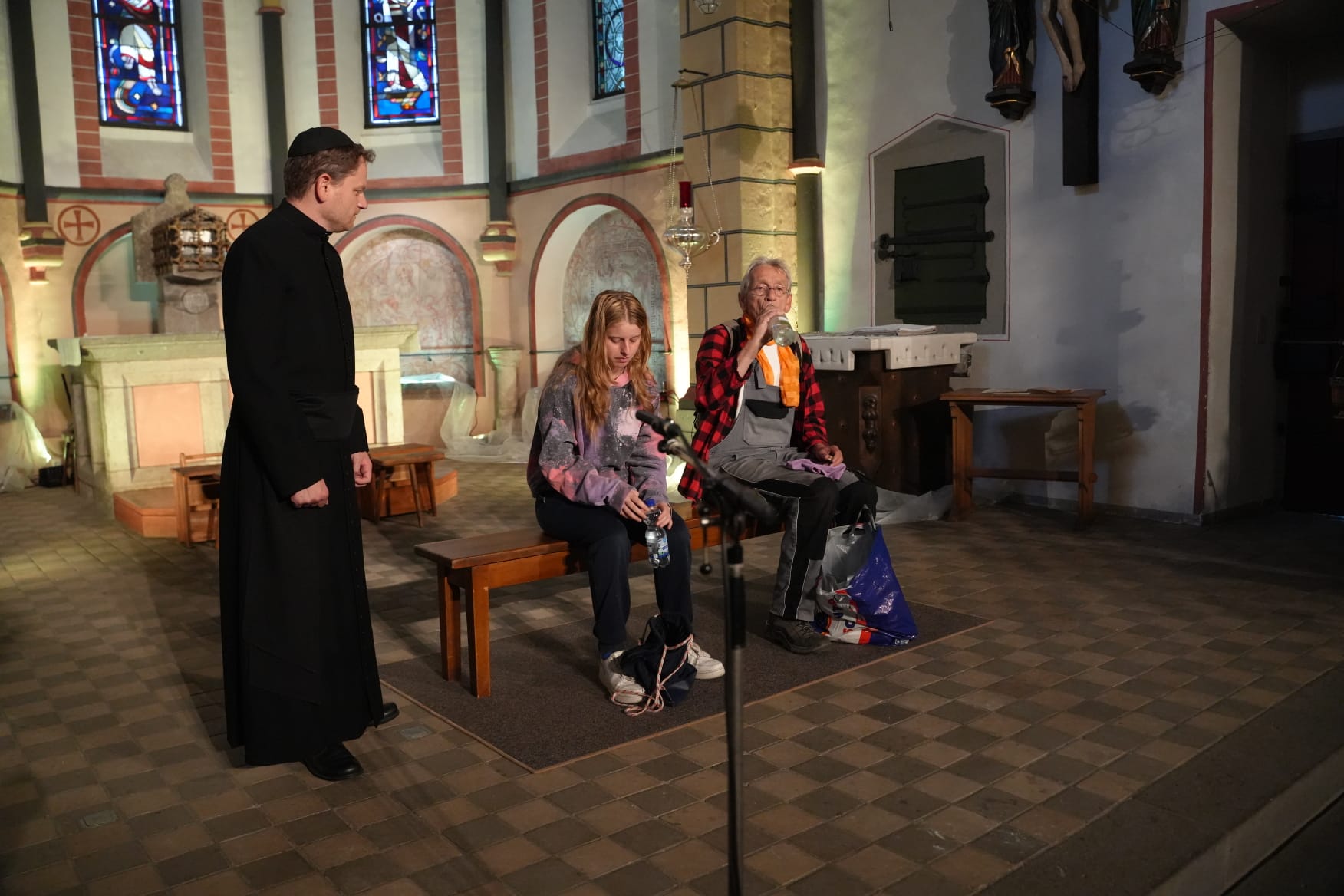 Eine Szene in einer Kirche: Zwei Personen sitzen auf einer Holzbank und wirken wie moderne Passanten oder Reisende mit Taschen und Rucksäcken. Eine weitere Person in langem schwarzem Gewand steht links neben ihnen. Der Hintergrund zeigt den Altarraum, Kirchenfenster und Steinboden.