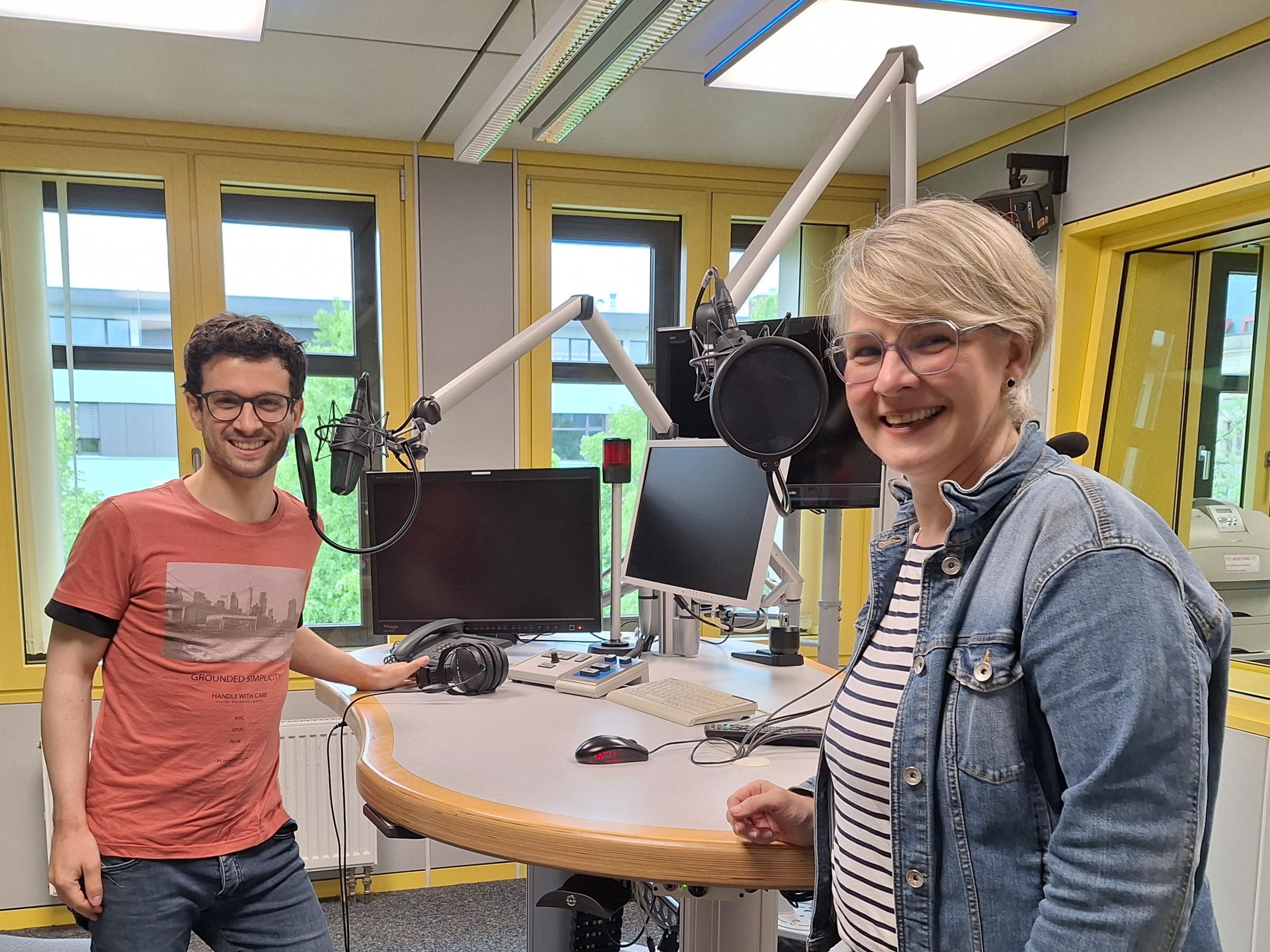 Das Foto zeigt Christopher Hoffmann (links) und Nicole Stockschlaeder (rechts). Sie stehen in einem Hörfunkstudio an einem Tisch mit Mikrofonen und lächeln freundlich in die Kamera.
