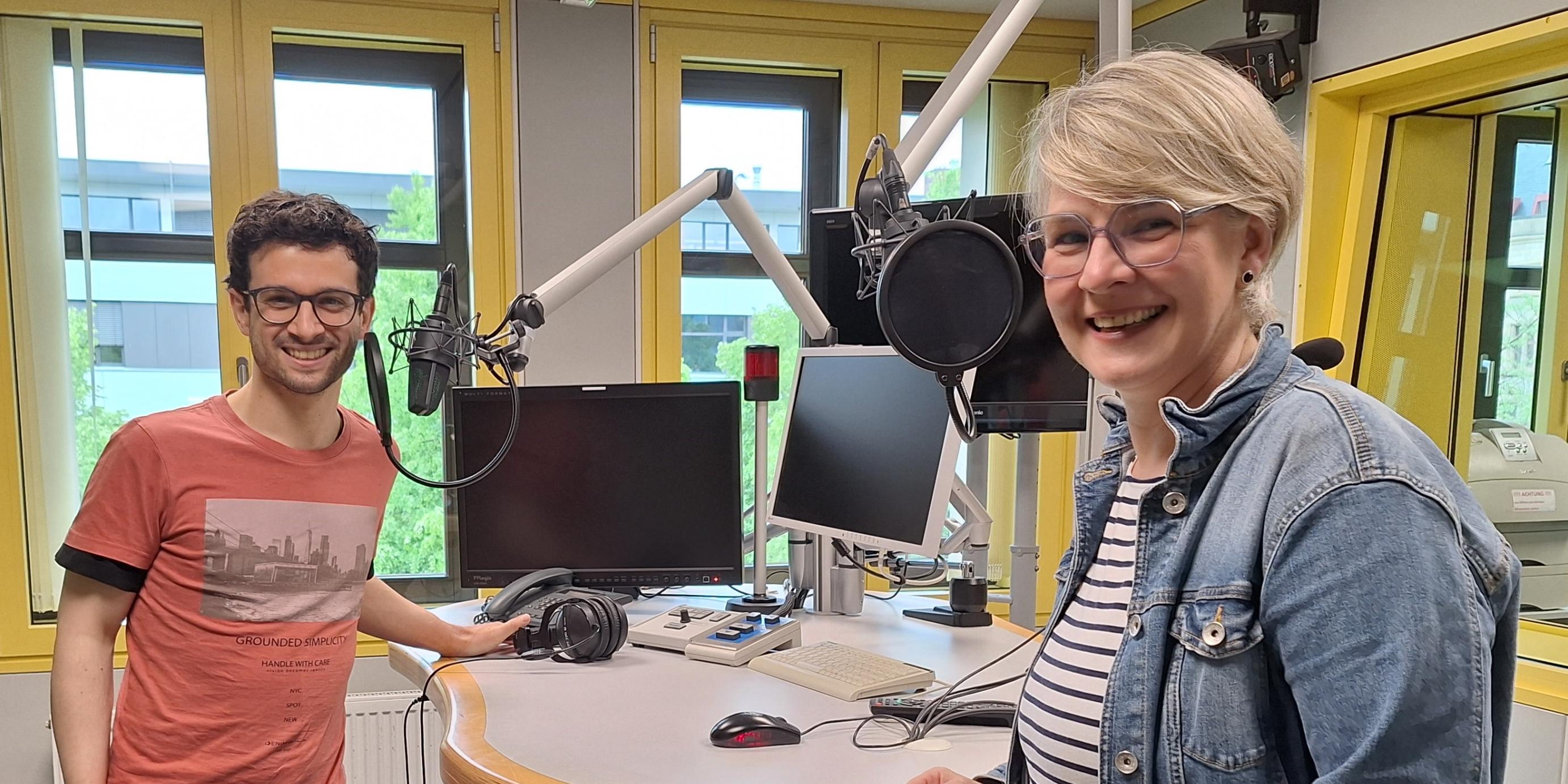 Das Foto zeigt Christopher Hoffmann (links) und Nicole Stockschlaeder (rechts). Sie stehen in einem Hörfunkstudio an einem Tisch mit Mikrofonen und lächeln freundlich in die Kamera.