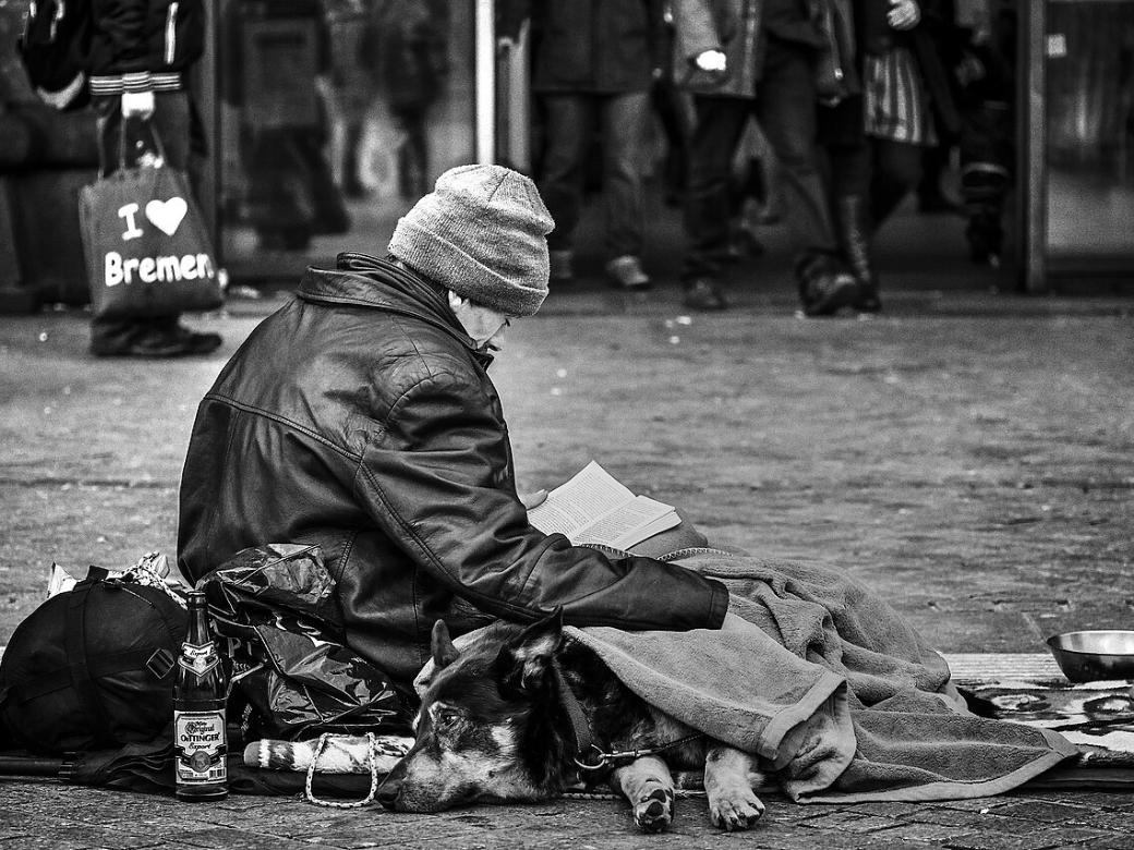 Obdachlose Person sitzt auf Straße, Hund liegt zugedeckt neben ihr.
