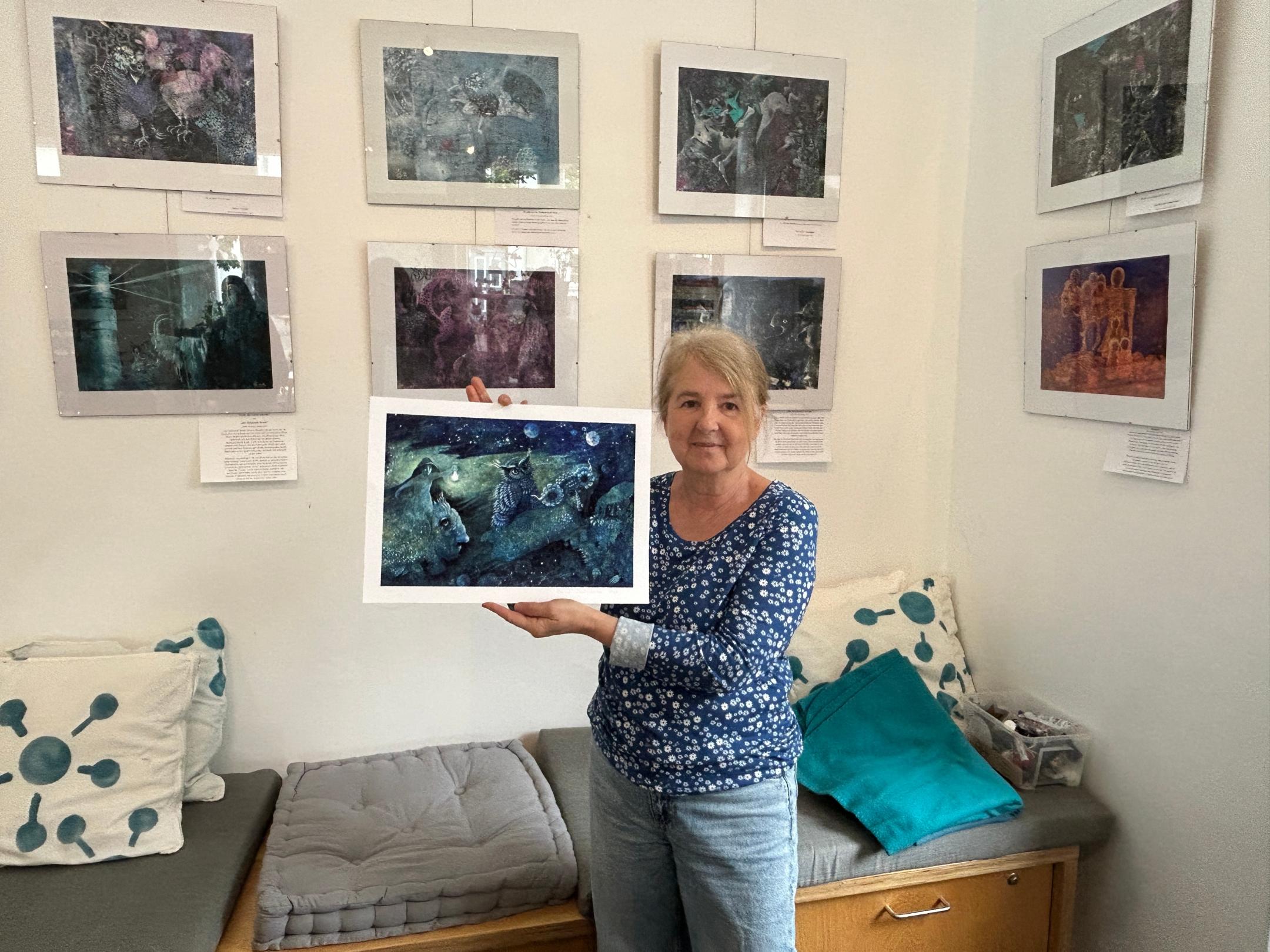 Eine Frau hält eins der von ihr gemalten Bilder in der Hand, weitere hängen im Hintergrund an der Wand.