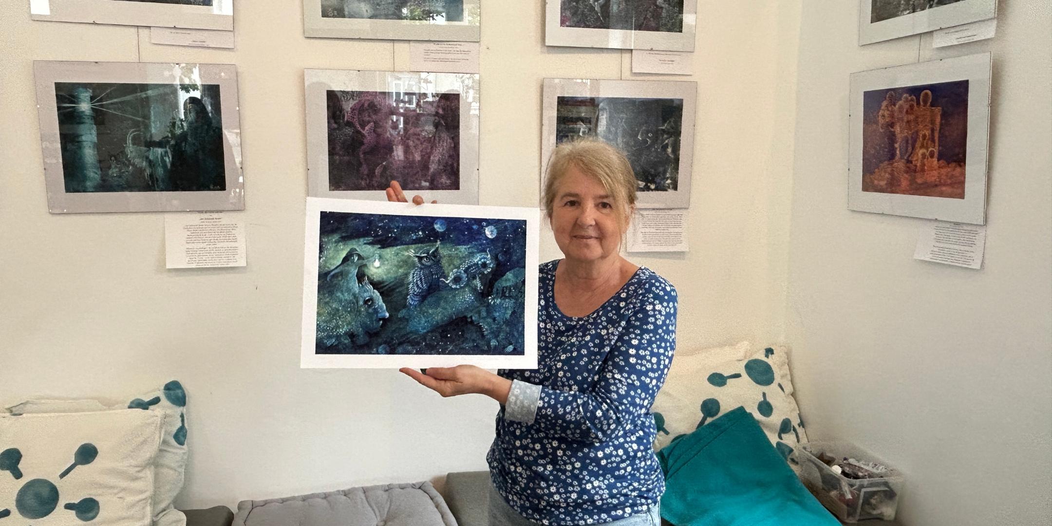 Eine Frau hält eins der von ihr gemalten Bilder in der Hand, weitere hängen im Hintergrund an der Wand.