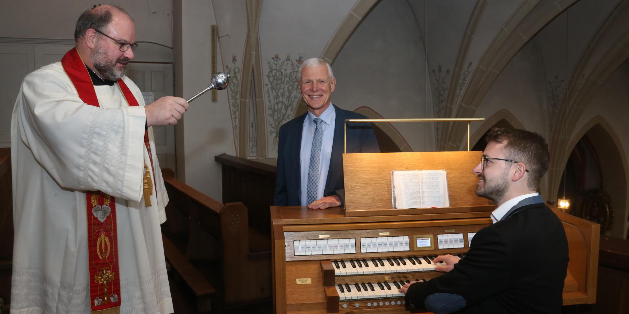 Ein Pfarrer besprengt eine Orgel mit Weihwasser, ein Mann sitzt an der Orgel, ein weiterer Mann steht hinter der Orgel und lächelt.