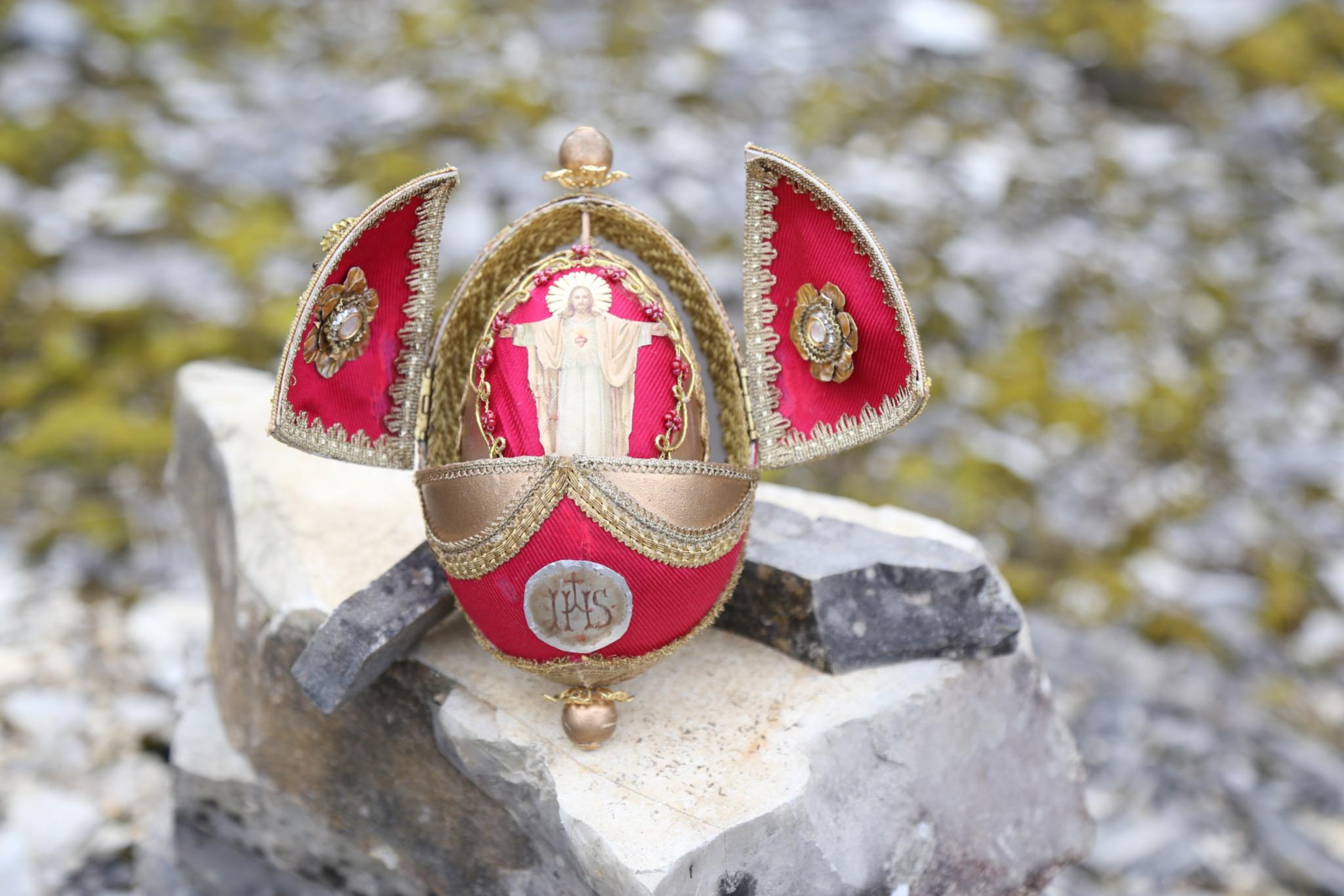 Ein verziertes, rot‑goldenes Osterei steht geöffnet auf einem großen Stein in einer felsigen Umgebung. Die beiden seitlichen Klappen des Eis sind nach außen aufgeklappt und mit goldenen Stickereien geschmückt. Im Inneren des Eis befindet sich eine kleine, hellfarbene Figur, die mit ausgebreiteten Armen dargestellt ist. Auf der Vorderseite des Eis ist ein rundes Emblem mit den Buchstaben ‚IHS‘ angebracht.