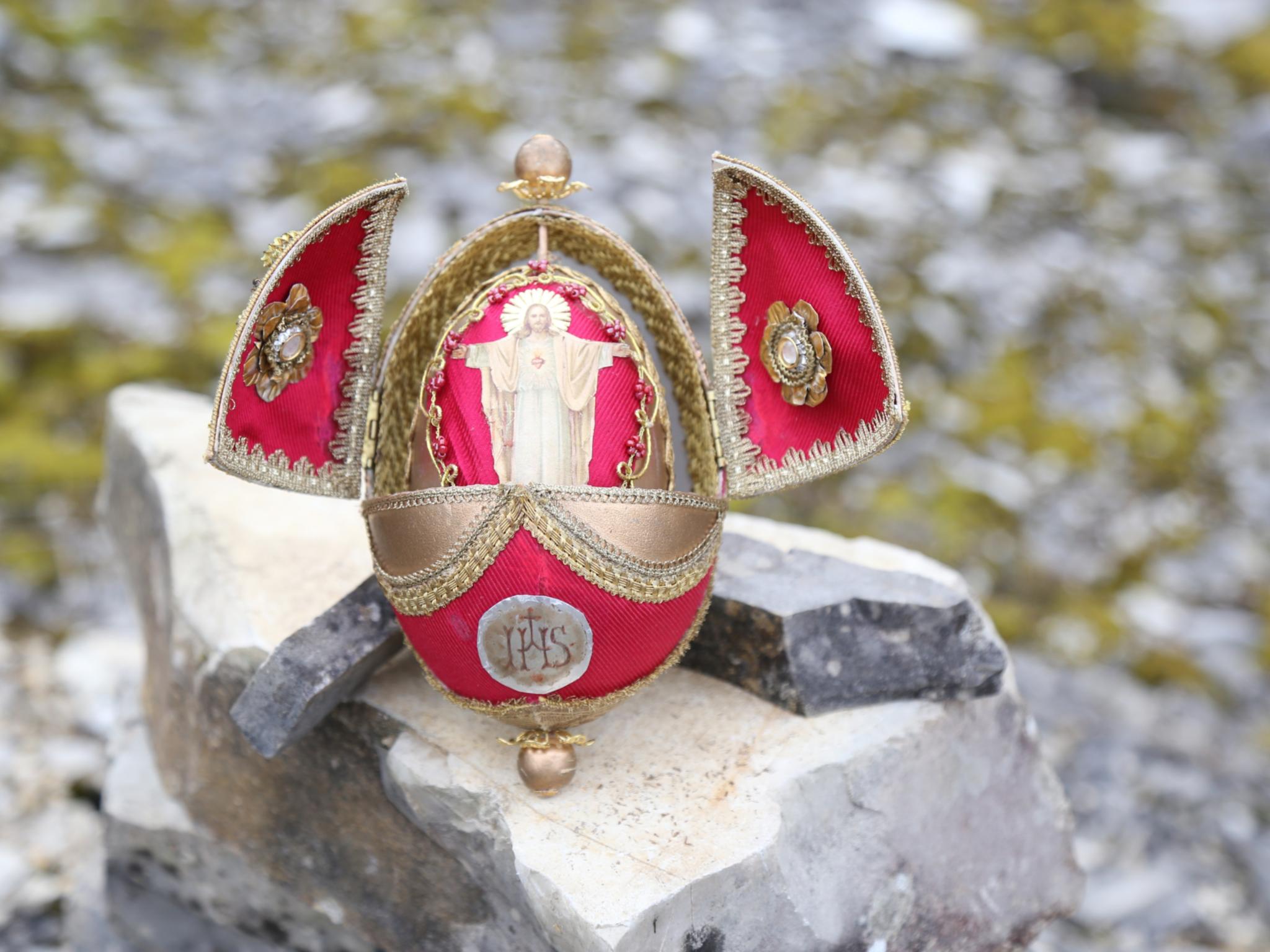Ein verziertes, rot‑goldenes Osterei steht geöffnet auf einem großen Stein in einer felsigen Umgebung. Die beiden seitlichen Klappen des Eis sind nach außen aufgeklappt und mit goldenen Stickereien geschmückt. Im Inneren des Eis befindet sich eine kleine, hellfarbene Figur, die mit ausgebreiteten Armen dargestellt ist. Auf der Vorderseite des Eis ist ein rundes Emblem mit den Buchstaben ‚IHS‘ angebracht.