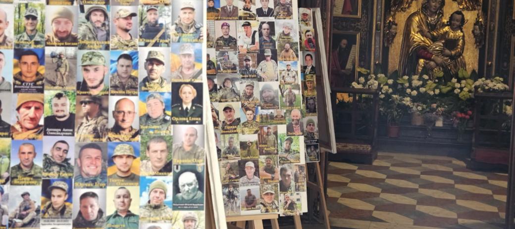 In einer Kirche stehen mehrere Staffeleien mit Fotos von Soldaten. Im Hintergrund befindet sich ein Altar mit einer goldenen Marienstatue und Blumenschmuck.