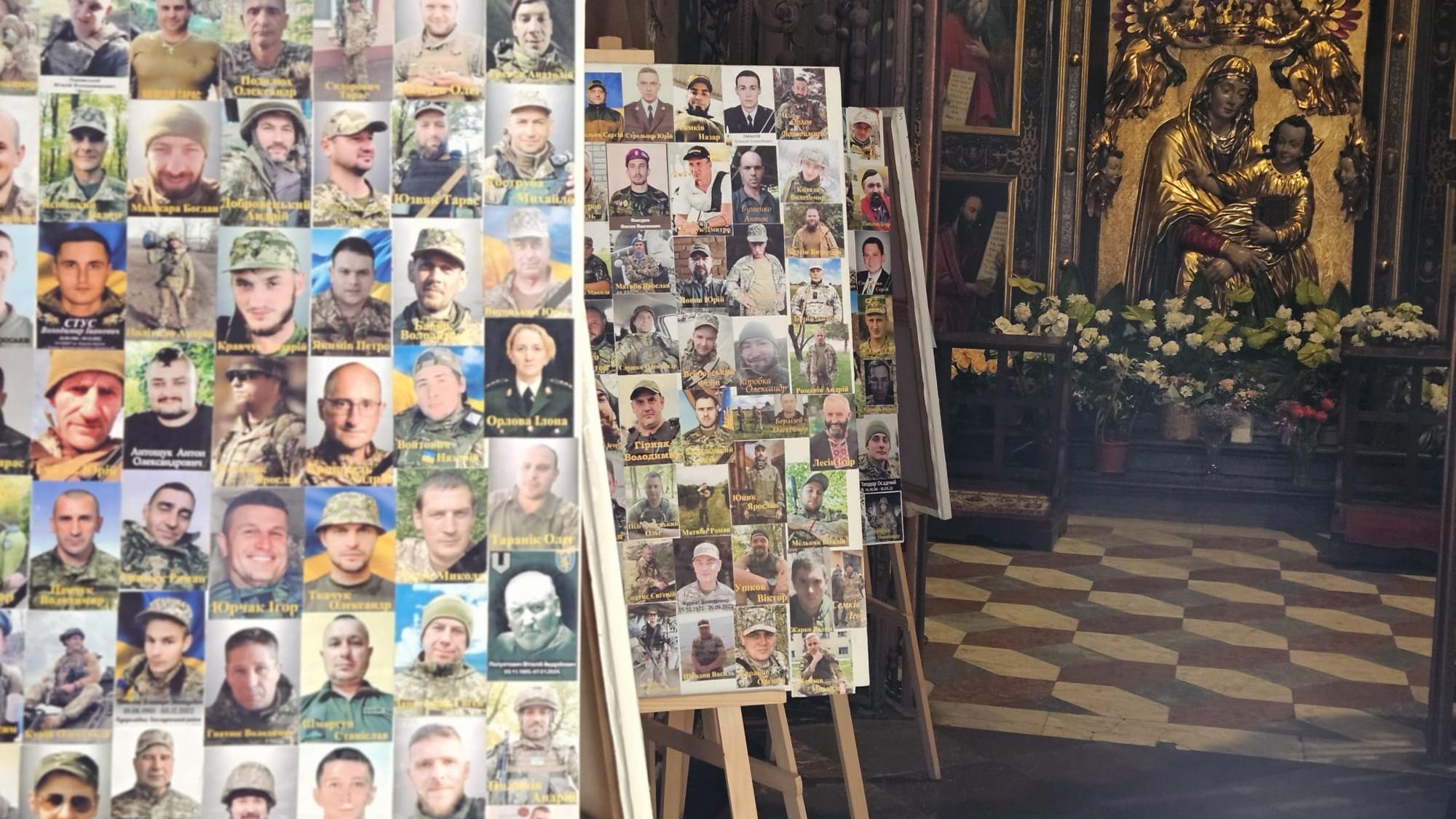 In einer Kirche stehen mehrere Staffeleien mit Fotos von Soldaten. Im Hintergrund befindet sich ein Altar mit einer goldenen Marienstatue und Blumenschmuck.