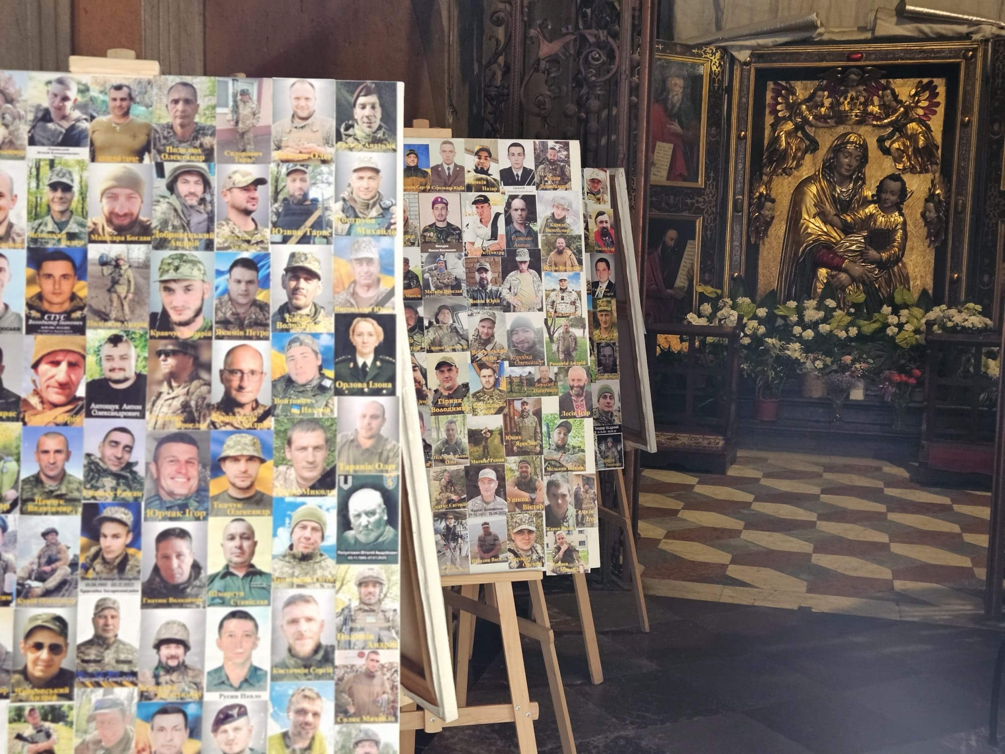In einer Kirche stehen mehrere Staffeleien mit Fotos von Soldaten. Im Hintergrund befindet sich ein Altar mit einer goldenen Marienstatue und Blumenschmuck.