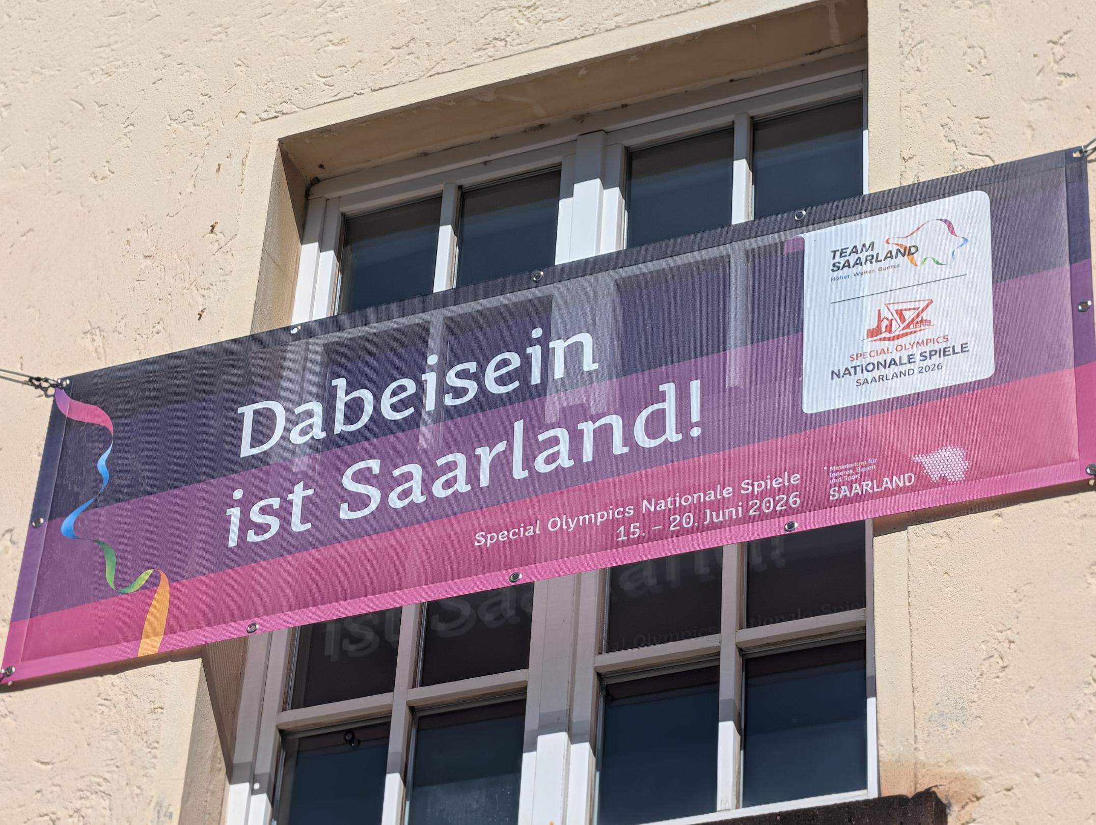 Ein Banner der Special Olympics Nationalen Spiele Saarland 2026 hängt vor den Fenstern eines hellen Gebäudes. Auf dem Banner steht in großen weißen Buchstaben: „Dabeisein ist Saarland!“. Rechts sind die Logos von Team Saarland und den Special Olympics zu sehen. Sonnenschein beleuchtet die Fassade.