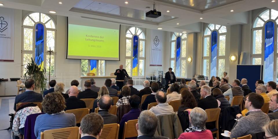 Man sieht viele Menschen, die in einem Saal sitzen und eine Präsentation anschauen