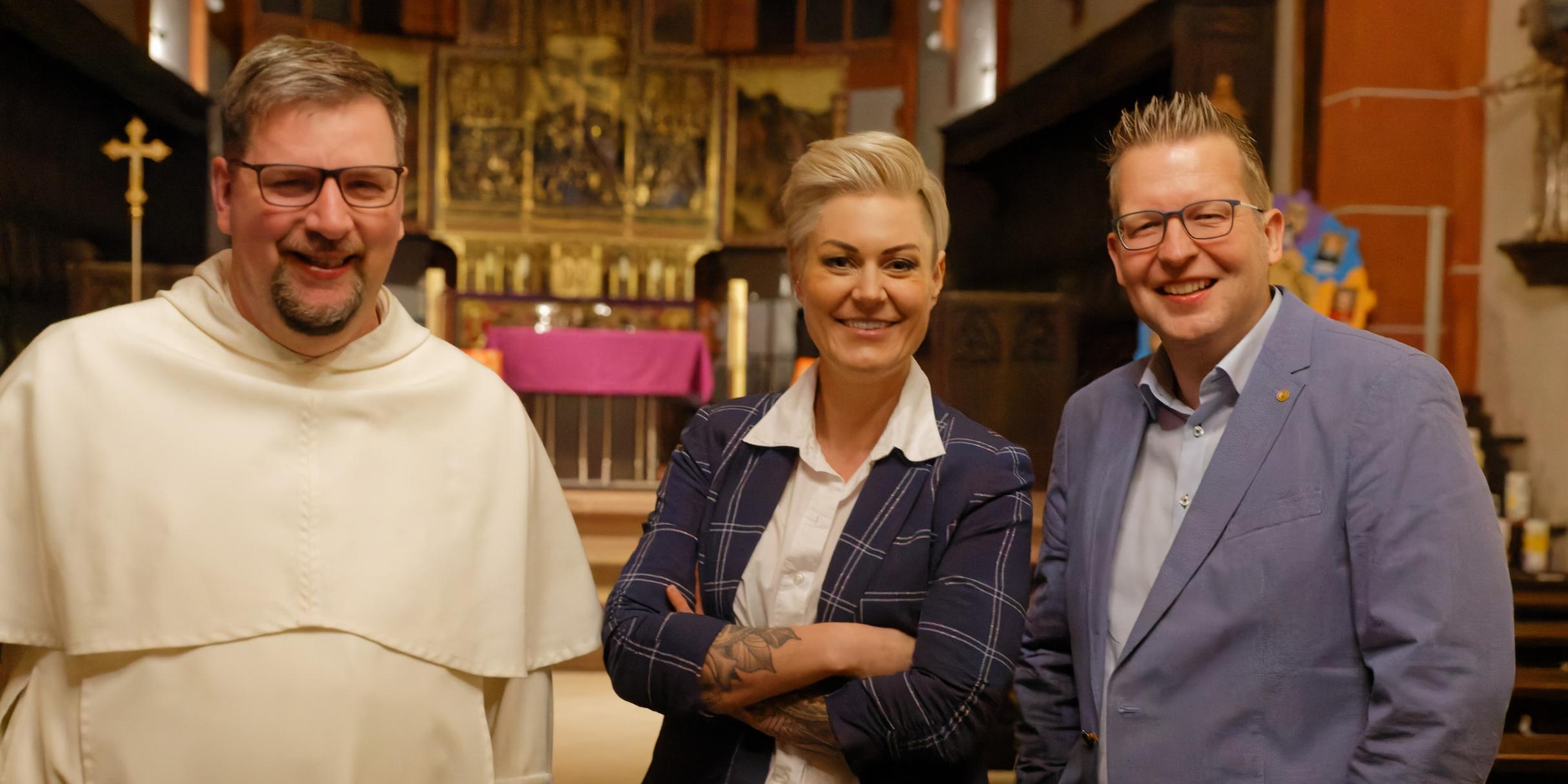 Das Foto zeigt drei Personen in einer Kirche, die freundlich in die Kamera lächeln. Von links nach rechts sind Pater Albert Seul in einer weißen Ordenskleidung, Dr. Carola Holzner aka Doc Caro in einem dunkelblauen karierten Blazer mit verschränkten Armen und Tobias Marenberg in einem hellblauen Anzug zu sehen. Der Hintergrund zeigt einen vergoldeten Hochaltar mit einer violetten Altardecke. (Alt-Text erstellt mit Unterstützung von ChatGPT)