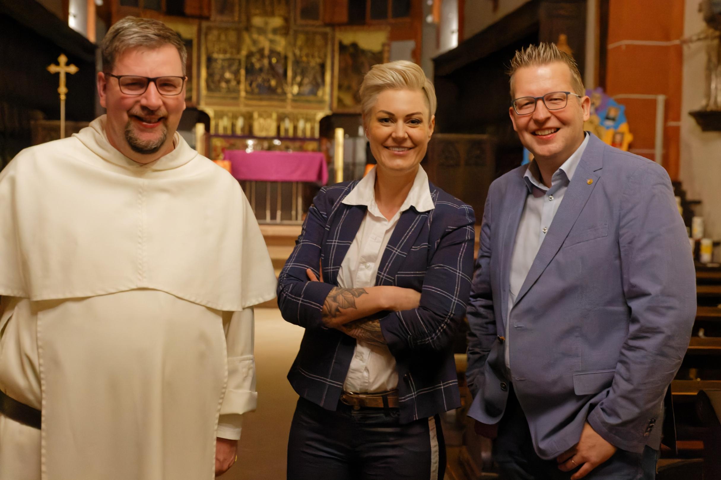 Das Foto zeigt drei Personen in einer Kirche, die freundlich in die Kamera lächeln. Von links nach rechts sind Pater Albert Seul in einer weißen Ordenskleidung, Dr. Carola Holzner aka Doc Caro in einem dunkelblauen karierten Blazer mit verschränkten Armen und Tobias Marenberg in einem hellblauen Anzug zu sehen. Der Hintergrund zeigt einen vergoldeten Hochaltar mit einer violetten Altardecke. (Alt-Text erstellt mit Unterstützung von ChatGPT)