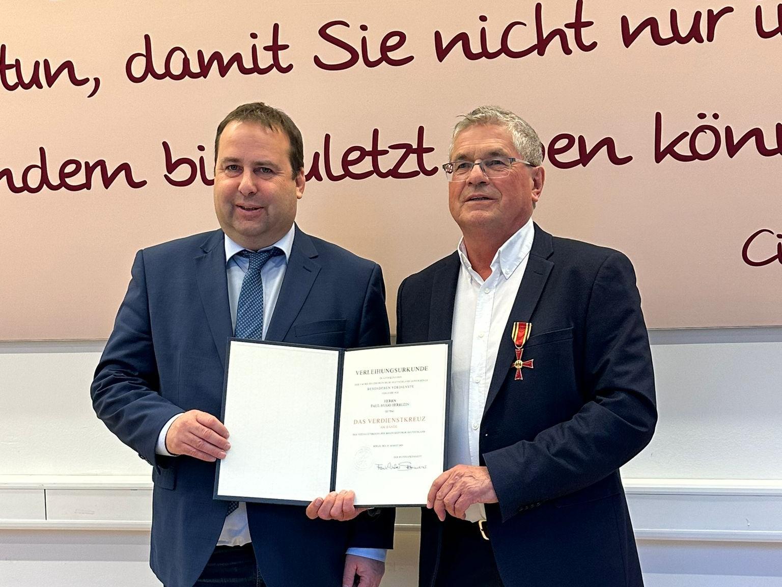 Zwei Personen stehen nebeneinander in einem Innenraum vor einer großen Wand mit dekorativer Schrift in dunkler Farbe. Beide tragen formelle Kleidung. Die Person links (Sozialminister Magnus Jung) hält eine geöffnete Urkunde mit beiden Händen. Die Person rechts (Paul Herrlein) trägt ein Jackett, an dem ein Orden mit rotem Band befestigt ist, und berührt die Urkunde mit einer Hand. Der Boden ist aus hellem Holz, und hinter den beiden verläuft ein weißes Paneel entlang der Wand.