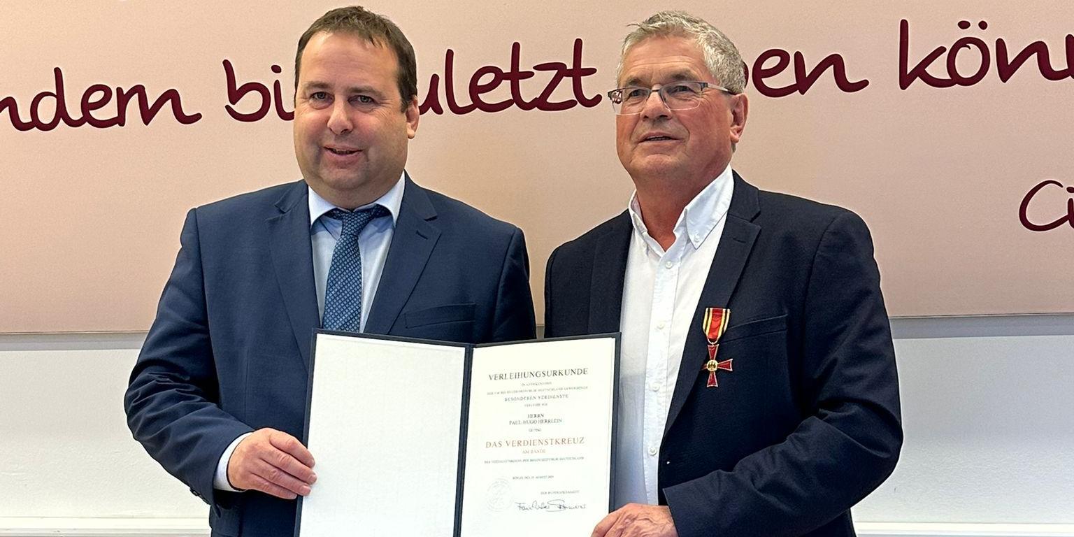 Zwei Personen stehen nebeneinander in einem Innenraum vor einer großen Wand mit dekorativer Schrift in dunkler Farbe. Beide tragen formelle Kleidung. Die Person links (Sozialminister Magnus Jung) hält eine geöffnete Urkunde mit beiden Händen. Die Person rechts (Paul Herrlein) trägt ein Jackett, an dem ein Orden mit rotem Band befestigt ist, und berührt die Urkunde mit einer Hand. Der Boden ist aus hellem Holz, und hinter den beiden verläuft ein weißes Paneel entlang der Wand.