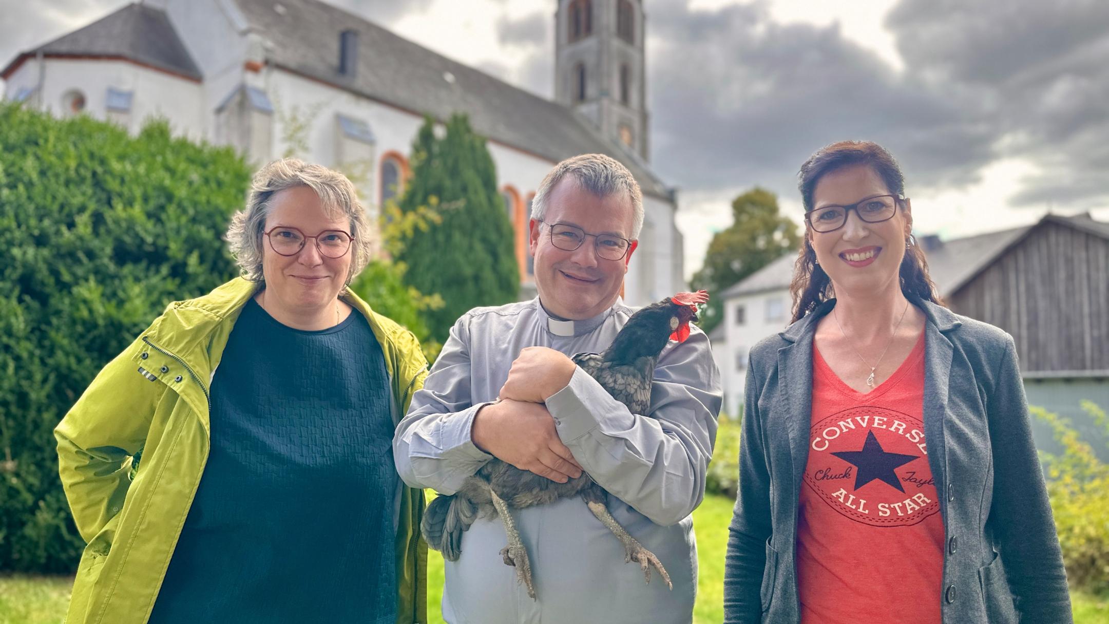 Gerlinde Paulus-Linn, Dekan Christian Heinz und Sandra Cäsar im Pfarrgarten mit Streichelzoo-Huhn Lieselotte.
