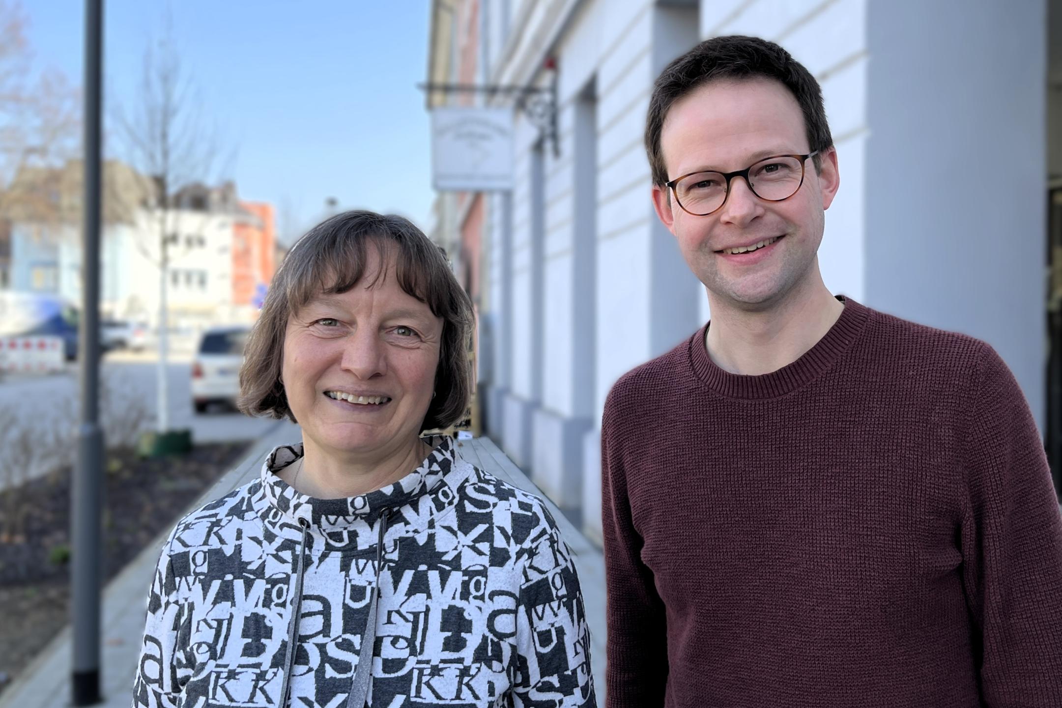 Eine Frau und ein junger Mann lachen in die Kamera und stehen in einer Einkaufsstraße.