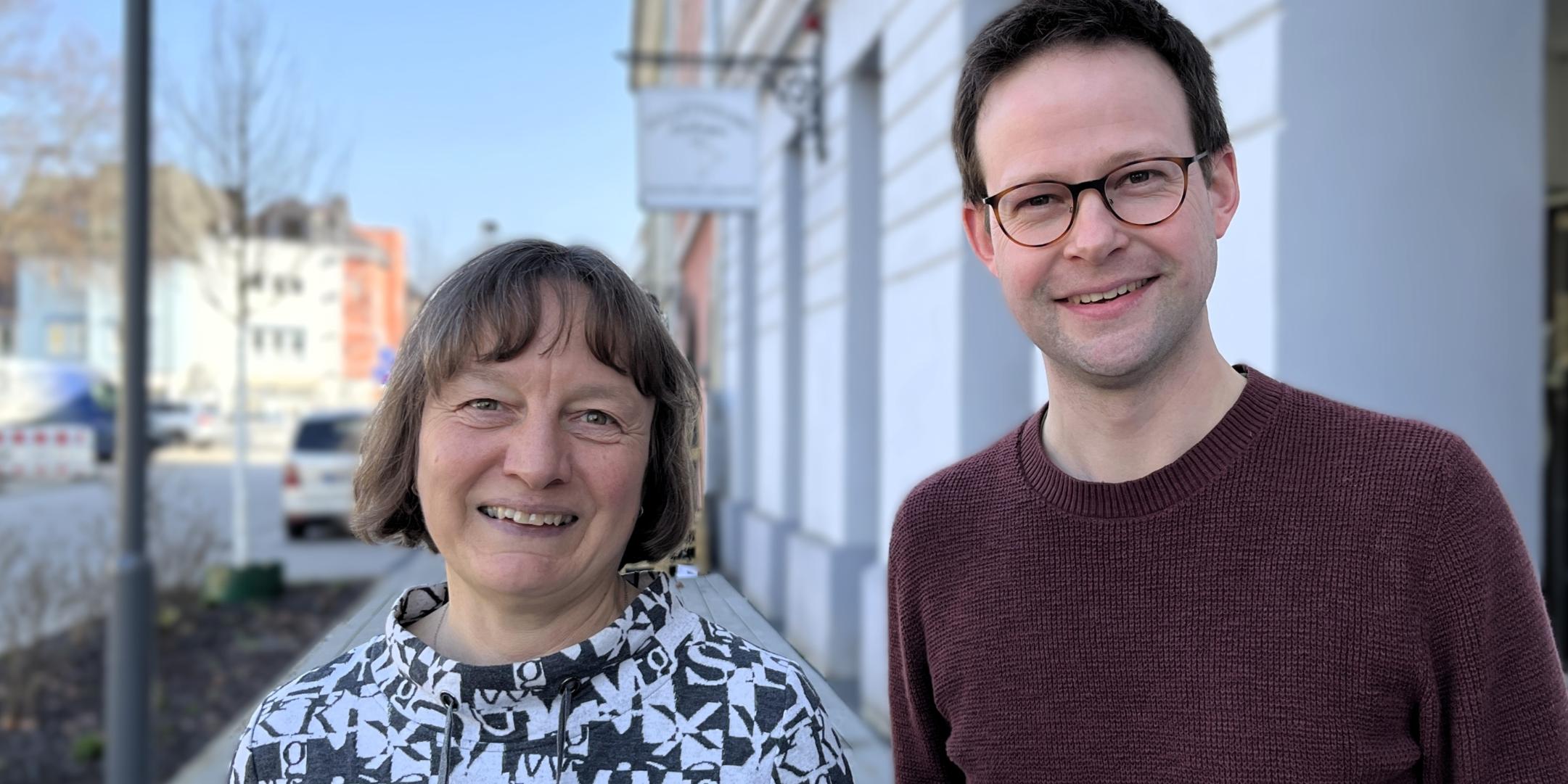 Eine Frau und ein junger Mann lachen in die Kamera und stehen in einer Einkaufsstraße.