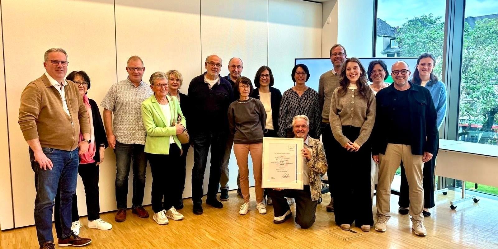 Ein Gruppenfoto mit unterschiedlichen Menschen, in der Mitte hält ein Mann eine Urkunde hoch.