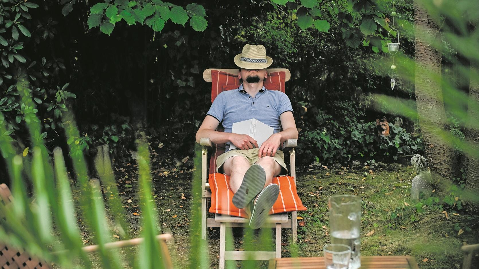 Ein Mann mit einem Hut auf dem Gesicht sitzt in einem Gartenstuhl und schläft, am 26. August 2025 in einem Garten in Bonn. Auf seinem Schoß liegt ein Buch.