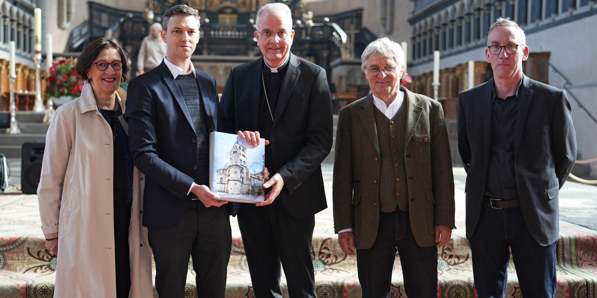 Vier Männer und eine Frau stehen im Trierer Dom vor dem Altar. Zwei Männer halten ein Buch in der Hand.