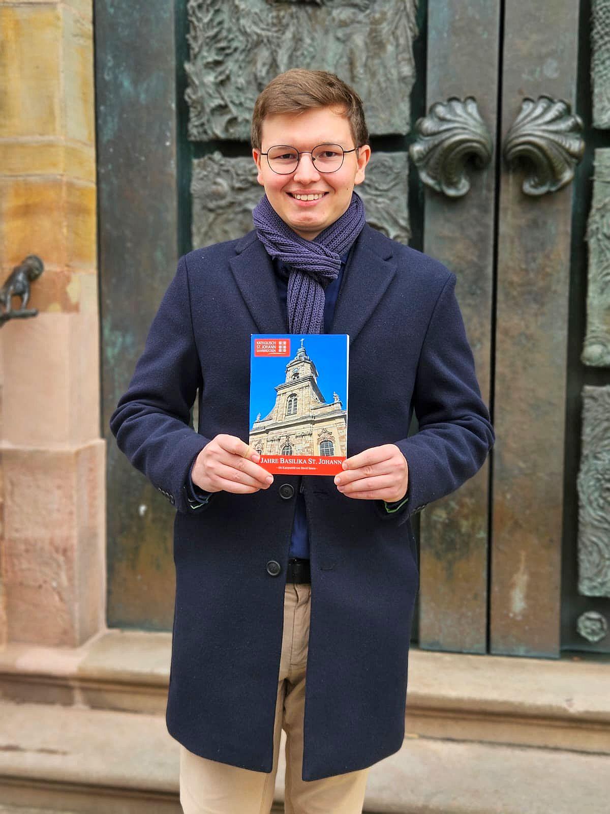 Der 19-jährige  David Sawas mit seiner Festschrift über die Saarbrücker Basilika St. Johann vor dem Portal der Kirche, das die künstlerische Handschrift des Bildhauers Ernst  Alt trägt.