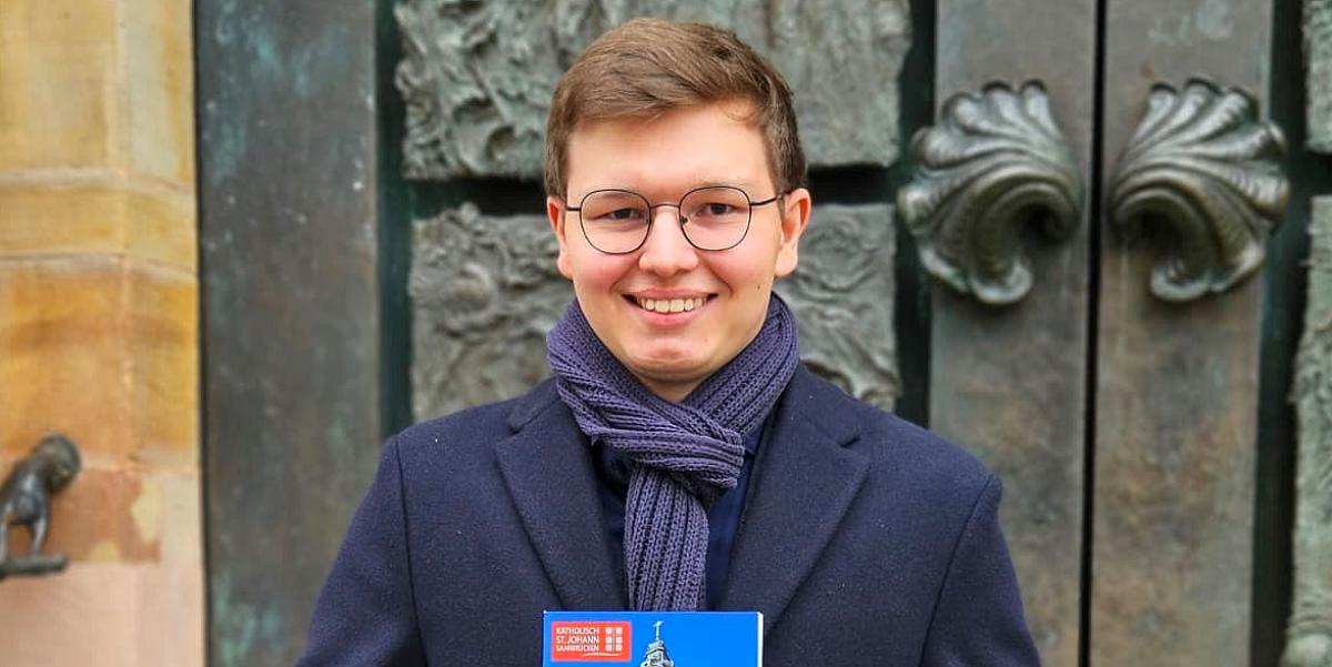 Der 19-jährige David Sawas mit seiner Festschrift über die Saarbrücker Basilika St. Johann vor dem Portal der Kirche, das die künstlerische Handschrift des Bildhauers Ernst Alt trägt.