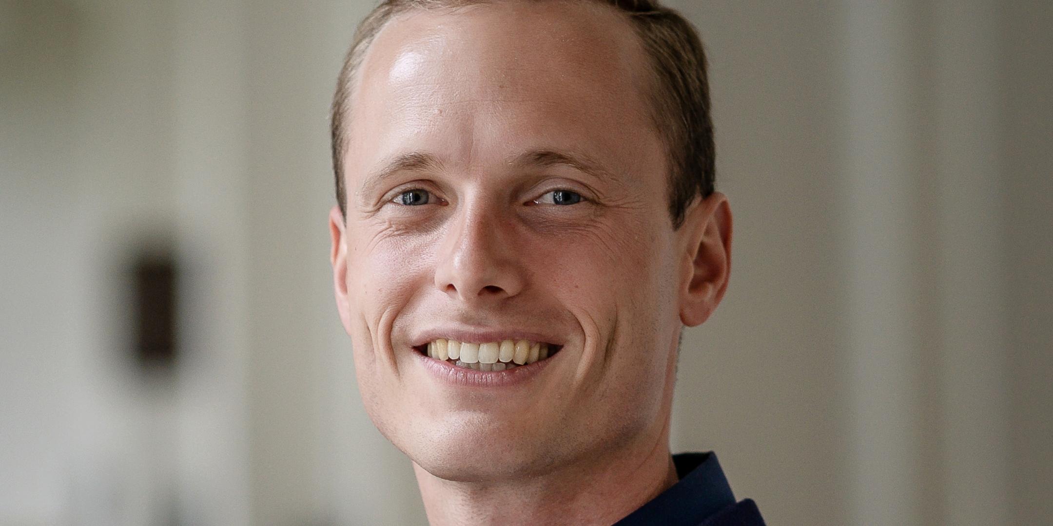 Mann in blauem Pullover steht in einem hellen Raum und lächelt in die Kamera