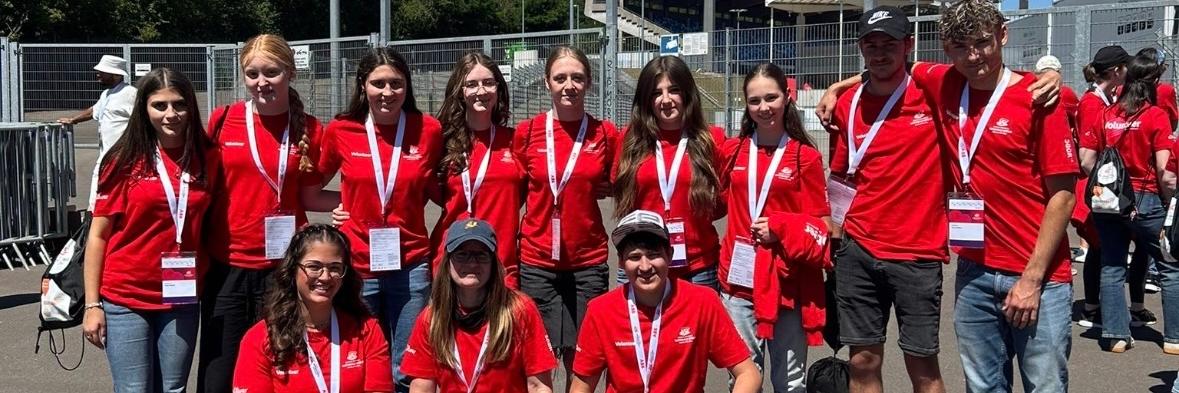 Gruppe von etwa elf Freiwilligen in roten T-Shirts mit Namensschildern und Akkreditierungen vor einem Stadioneingang. Die Personen stehen und knien für ein Gruppenfoto auf einem asphaltierten Platz. Im Hintergrund sind Zäune, Tribünen, Flutlichtmasten und orangefarbene Schilder mit den Aufschriften „Zufahrt Gäste-Fanbusse“ und „Zugang Gästefans“ zu sehen. Sonniger Tag mit klarem blauem Himmel.