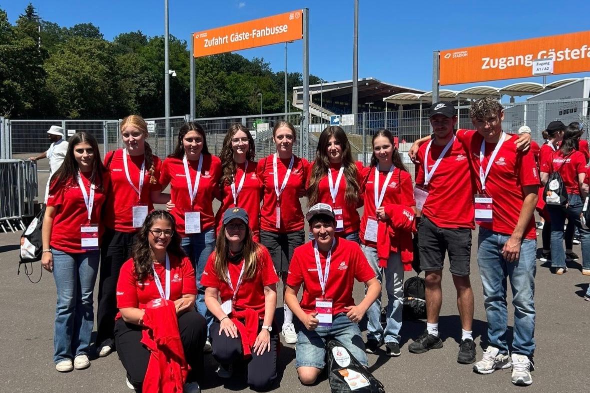 Gruppe von etwa elf Freiwilligen in roten T-Shirts mit Namensschildern und Akkreditierungen vor einem Stadioneingang. Die Personen stehen und knien für ein Gruppenfoto auf einem asphaltierten Platz. Im Hintergrund sind Zäune, Tribünen, Flutlichtmasten und orangefarbene Schilder mit den Aufschriften „Zufahrt Gäste-Fanbusse“ und „Zugang Gästefans“ zu sehen. Sonniger Tag mit klarem blauem Himmel.