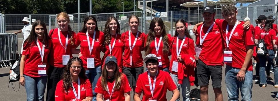 Gruppe von etwa elf Freiwilligen in roten T-Shirts mit Namensschildern und Akkreditierungen vor einem Stadioneingang. Die Personen stehen und knien für ein Gruppenfoto auf einem asphaltierten Platz. Im Hintergrund sind Zäune, Tribünen, Flutlichtmasten und orangefarbene Schilder mit den Aufschriften „Zufahrt Gäste-Fanbusse“ und „Zugang Gästefans“ zu sehen. Sonniger Tag mit klarem blauem Himmel.
