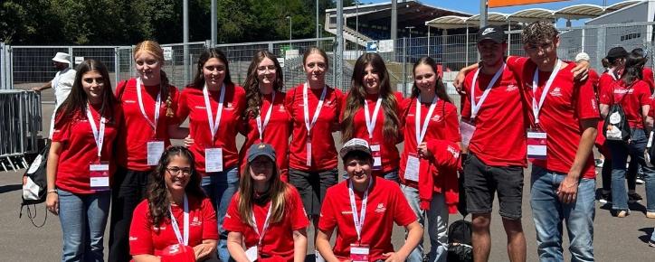 Gruppe von etwa elf Freiwilligen in roten T-Shirts mit Namensschildern und Akkreditierungen vor einem Stadioneingang. Die Personen stehen und knien für ein Gruppenfoto auf einem asphaltierten Platz. Im Hintergrund sind Zäune, Tribünen, Flutlichtmasten und orangefarbene Schilder mit den Aufschriften „Zufahrt Gäste-Fanbusse“ und „Zugang Gästefans“ zu sehen. Sonniger Tag mit klarem blauem Himmel.