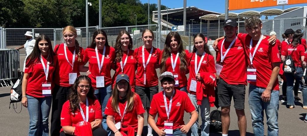 Gruppe von etwa elf Freiwilligen in roten T-Shirts mit Namensschildern und Akkreditierungen vor einem Stadioneingang. Die Personen stehen und knien für ein Gruppenfoto auf einem asphaltierten Platz. Im Hintergrund sind Zäune, Tribünen, Flutlichtmasten und orangefarbene Schilder mit den Aufschriften „Zufahrt Gäste-Fanbusse“ und „Zugang Gästefans“ zu sehen. Sonniger Tag mit klarem blauem Himmel.
