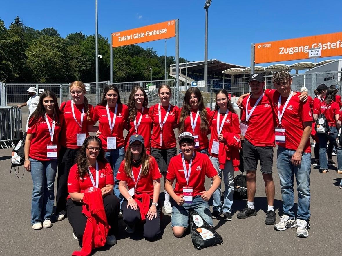 Gruppe von etwa elf Freiwilligen in roten T-Shirts mit Namensschildern und Akkreditierungen vor einem Stadioneingang. Die Personen stehen und knien für ein Gruppenfoto auf einem asphaltierten Platz. Im Hintergrund sind Zäune, Tribünen, Flutlichtmasten und orangefarbene Schilder mit den Aufschriften „Zufahrt Gäste-Fanbusse“ und „Zugang Gästefans“ zu sehen. Sonniger Tag mit klarem blauem Himmel.